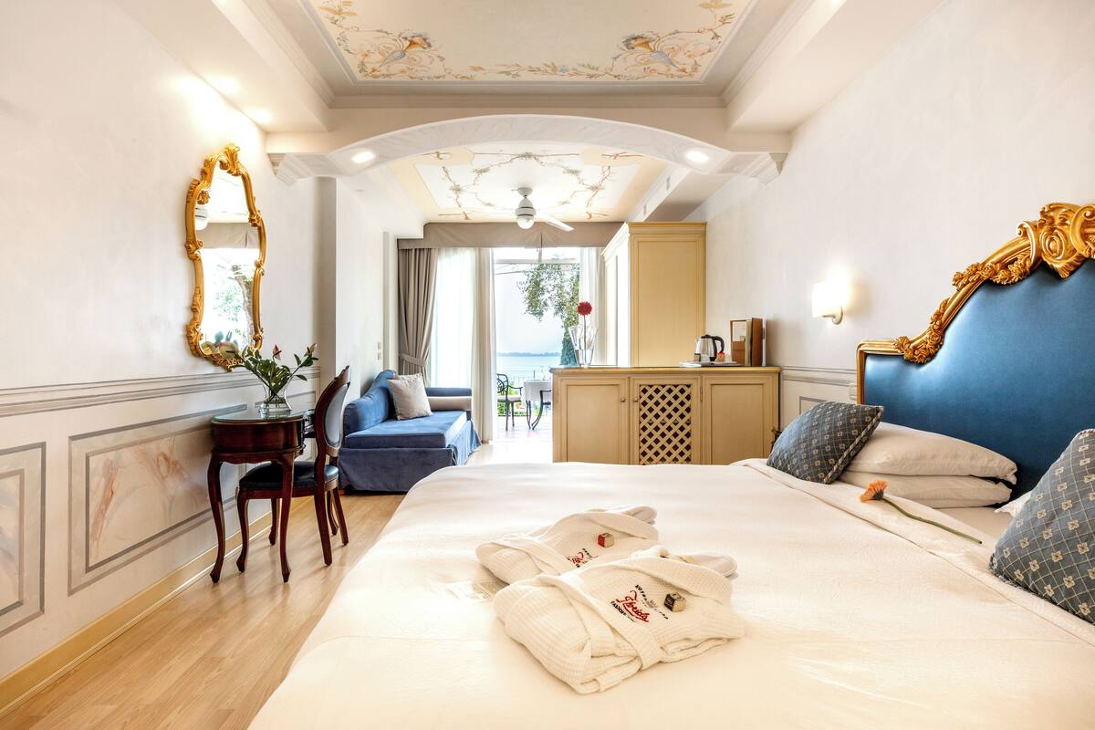 A hotel room with a large bed, a blue headboard, a wooden nightstand, a desk with a chair, and a view of the outdoors.