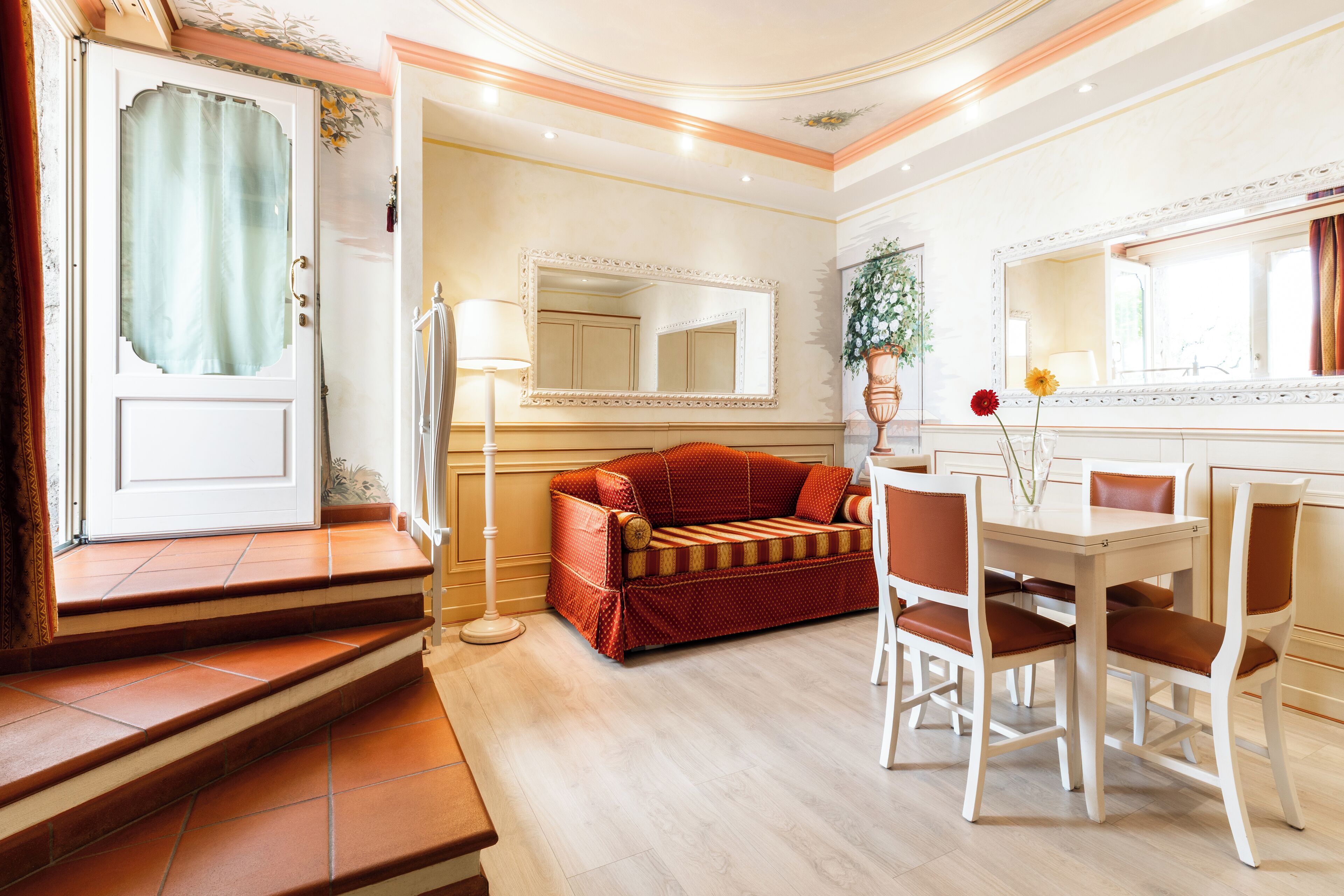 A hotel room with a sofa, dining table, chairs, a mirror, and a staircase.