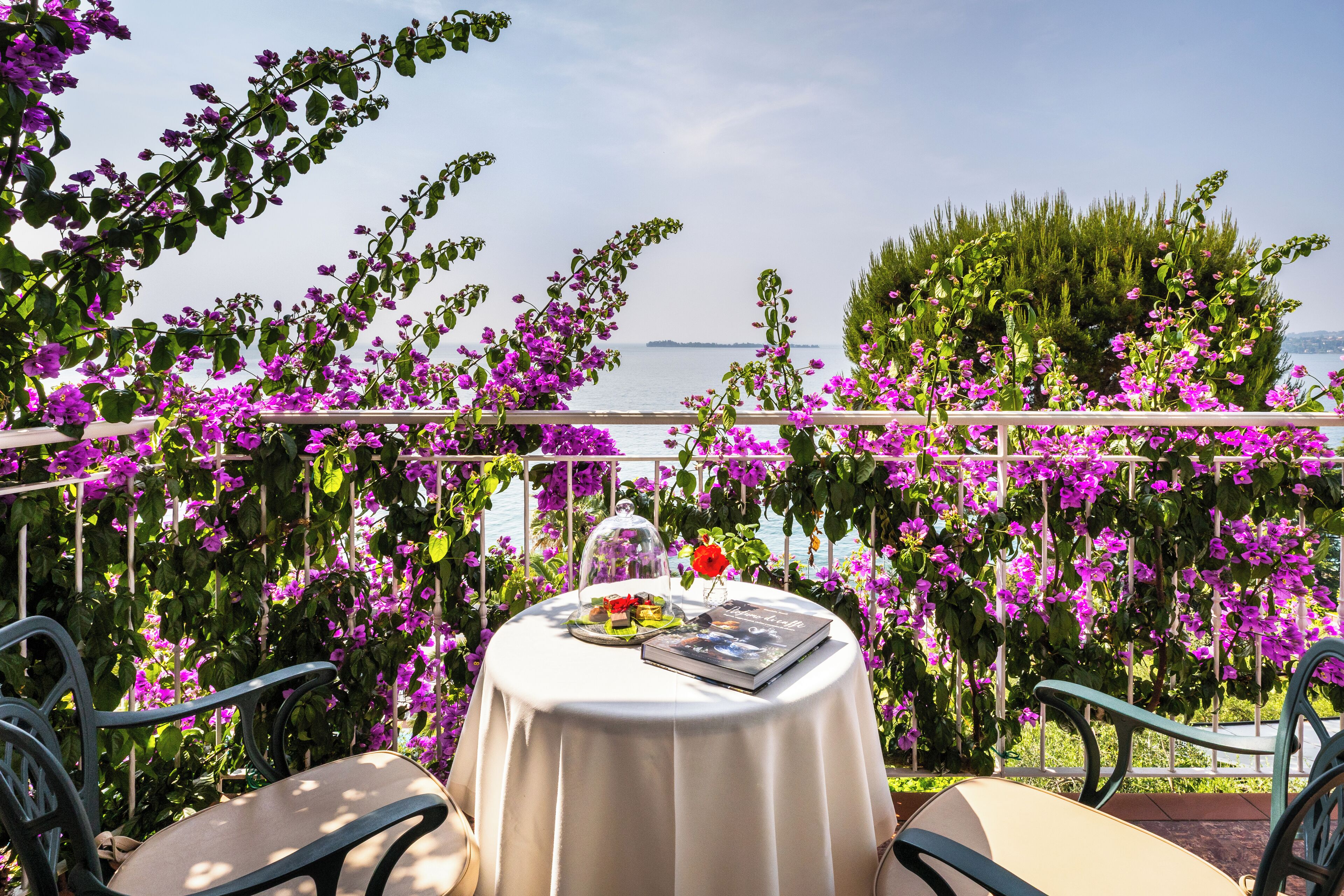 Un tavolo rotondo con tovaglia bianca, circondato da sedie, con sullo sfondo fiori viola e vista mare.