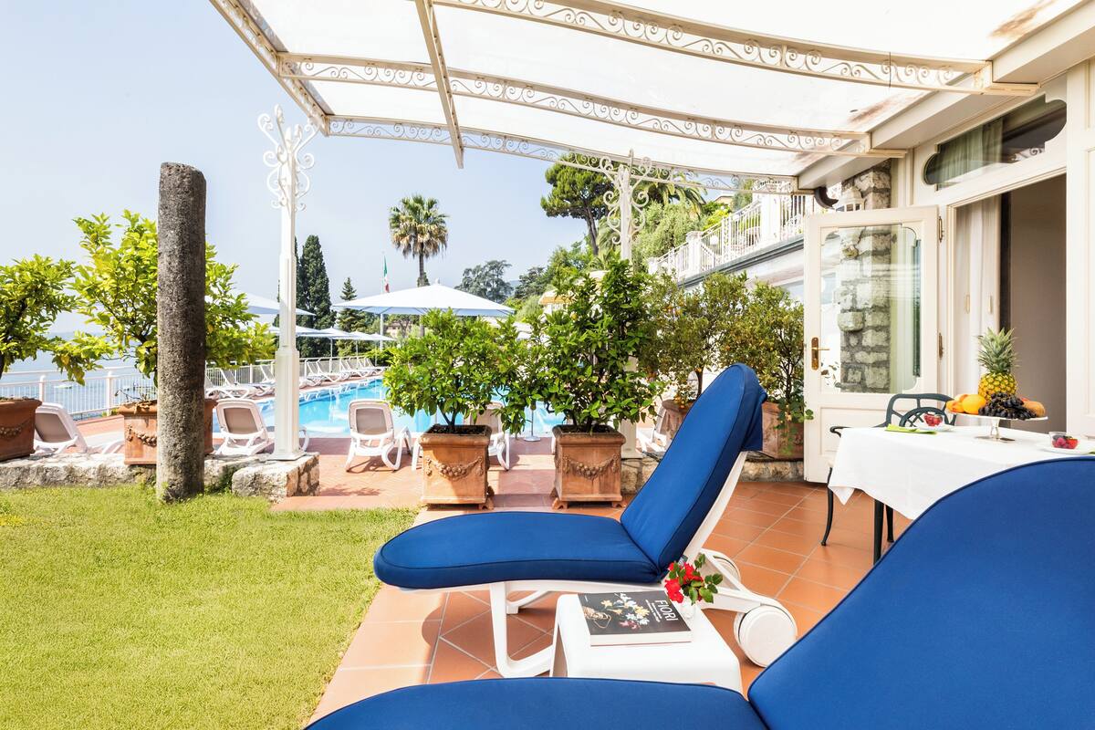 A patio area with two blue lounge chairs, a table with a book and a flower, and a view of a swimming pool and greenery.
