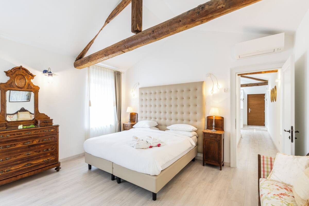 A bedroom with a large bed, wooden dresser, and a mirror. There is a door leading to another room.