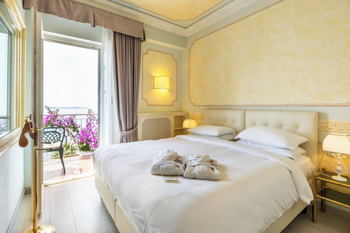 A hotel room with a large bed, a nightstand with a lamp, a view of the outdoors, and a balcony with flowers.