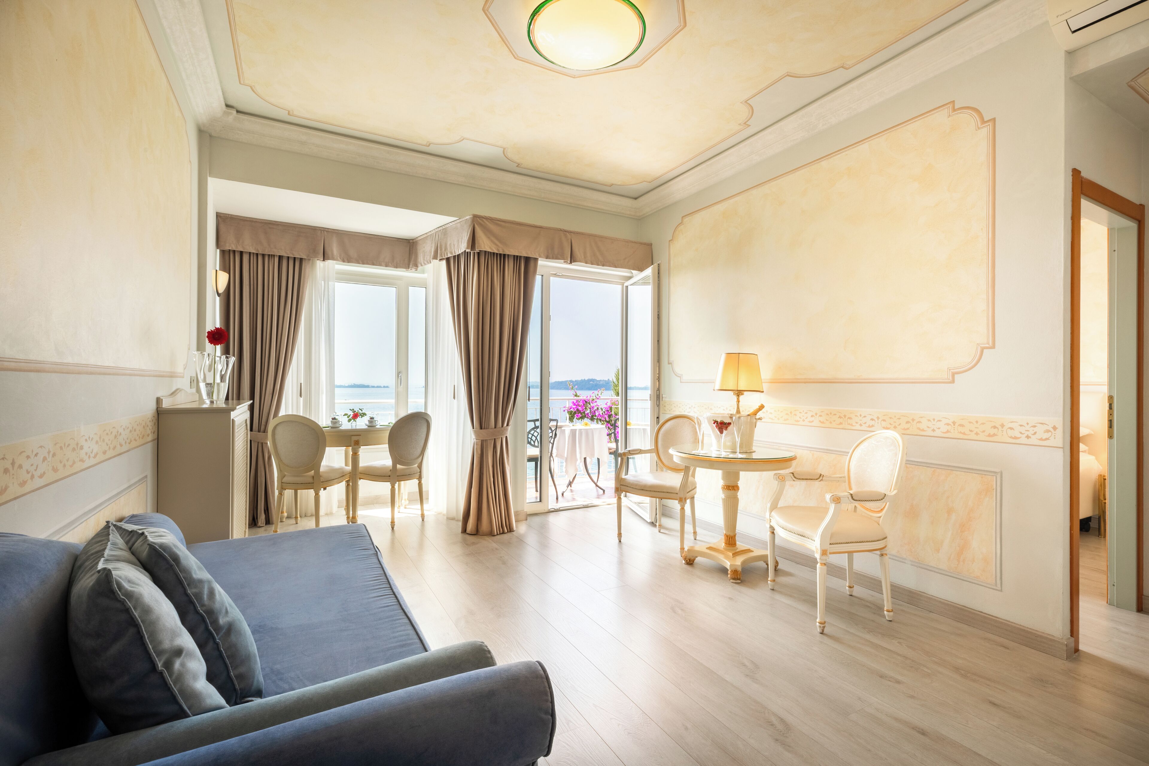 A hotel room with a sofa, a dining table with chairs, a lamp, and a view of the outdoors.