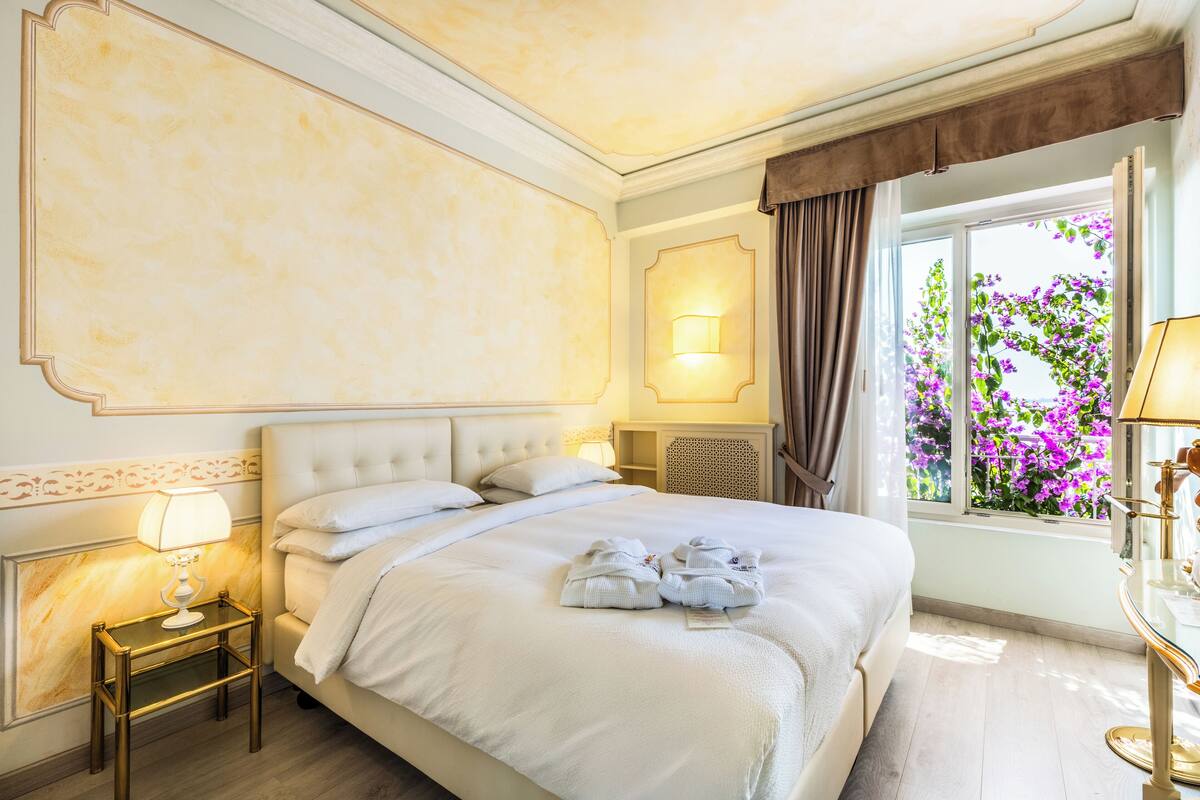 A hotel room with a large bed, a nightstand with a lamp, a window with curtains, and a view of purple flowers outside.