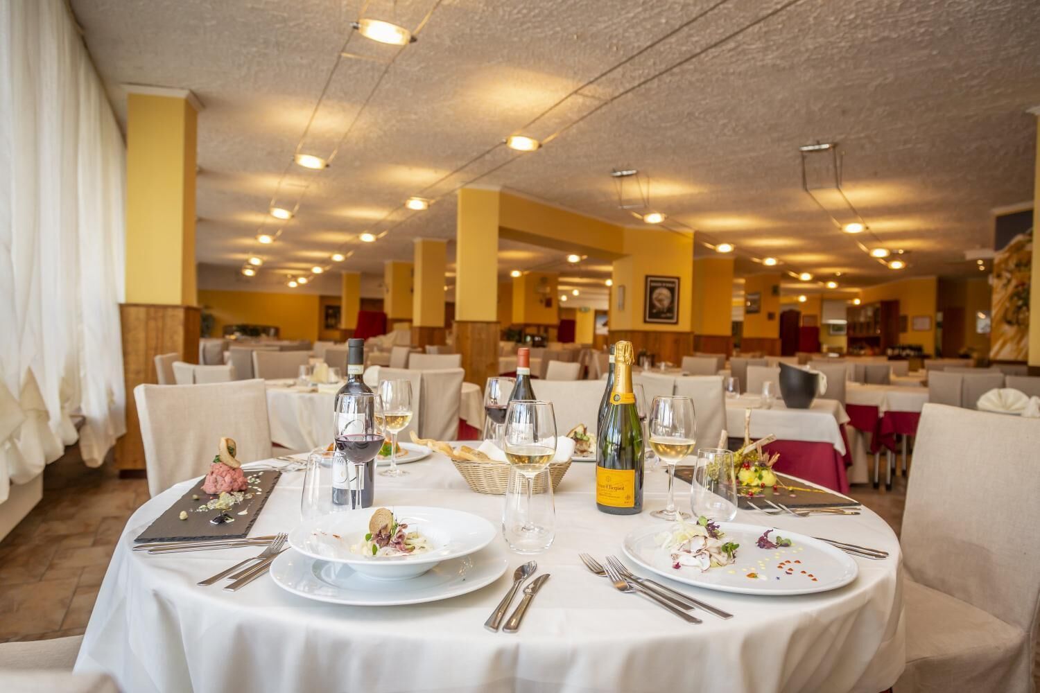 Restaurante com mesas redondas preparadas para refeições, com toalhas brancas, copos de vinho e garrafas.