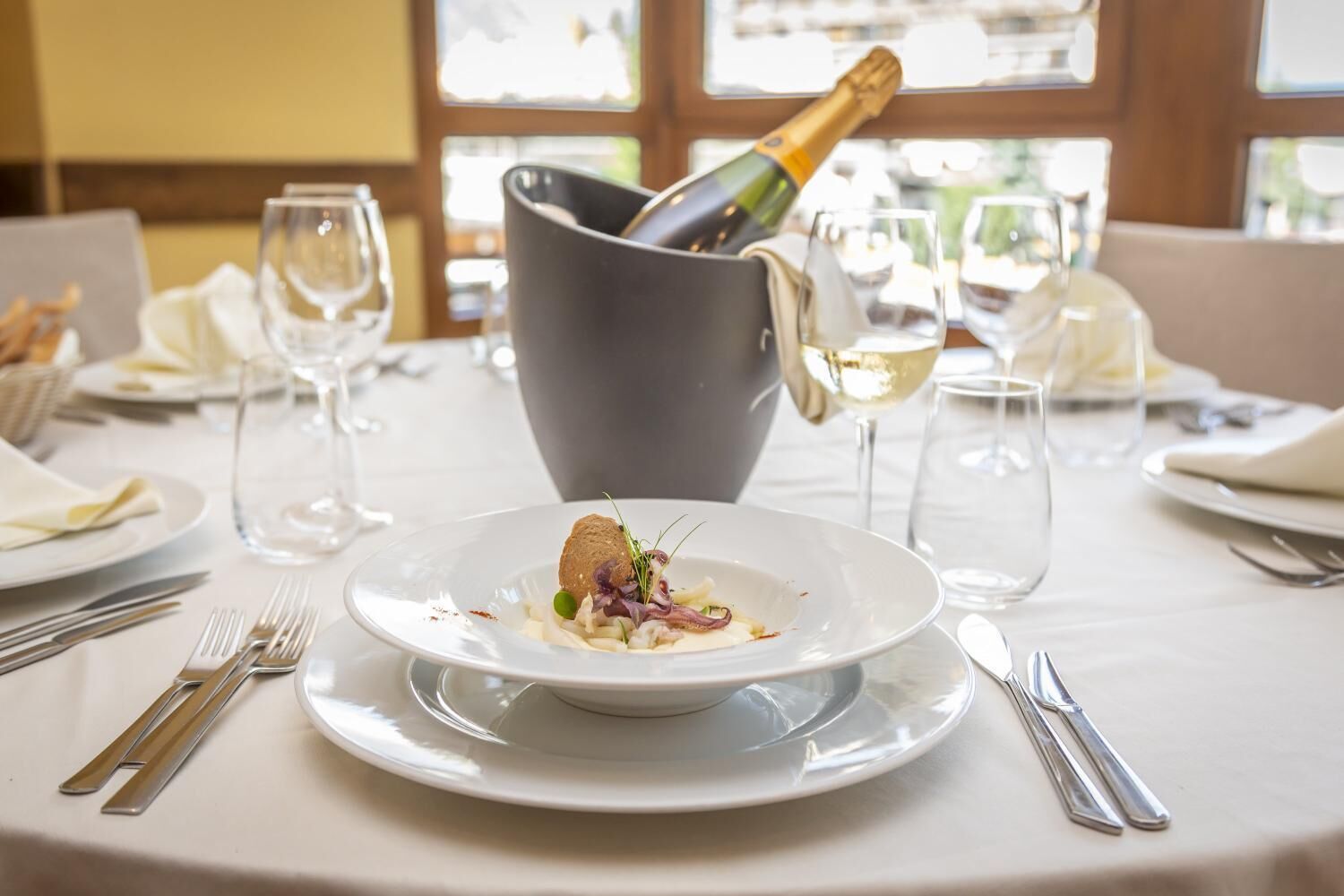 A dining table set with a white tablecloth, a plate of food, wine glasses, and a bottle of champagne.