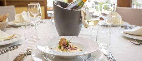 A dining table set with a white tablecloth, a plate of food, wine glasses, and a bottle of champagne.
