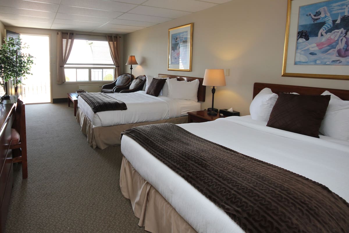 Quarto de hotel com duas camas, uma escrivaninha, cadeiras, uma janela e um quadro na parede.
