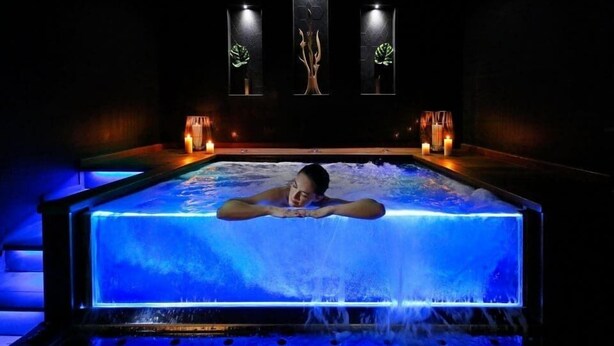 Une personne se trouve dans un grand bain à remous éclairé, doté d’une cascade artificielle, et est entourée de bougies et de panneaux décoratifs.
