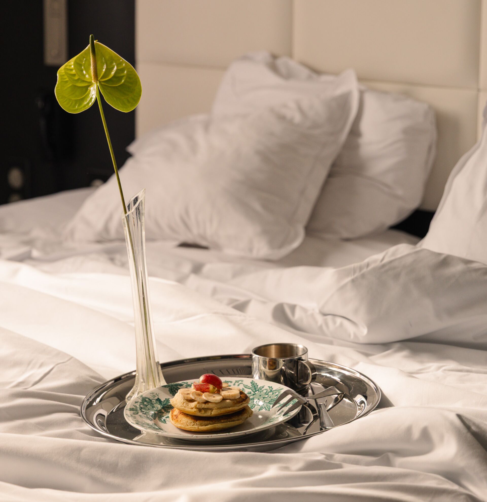 A neatly made bed with white linens, a tray with a plate of pancakes and a small cup, and a tall vase with a single green leaf.