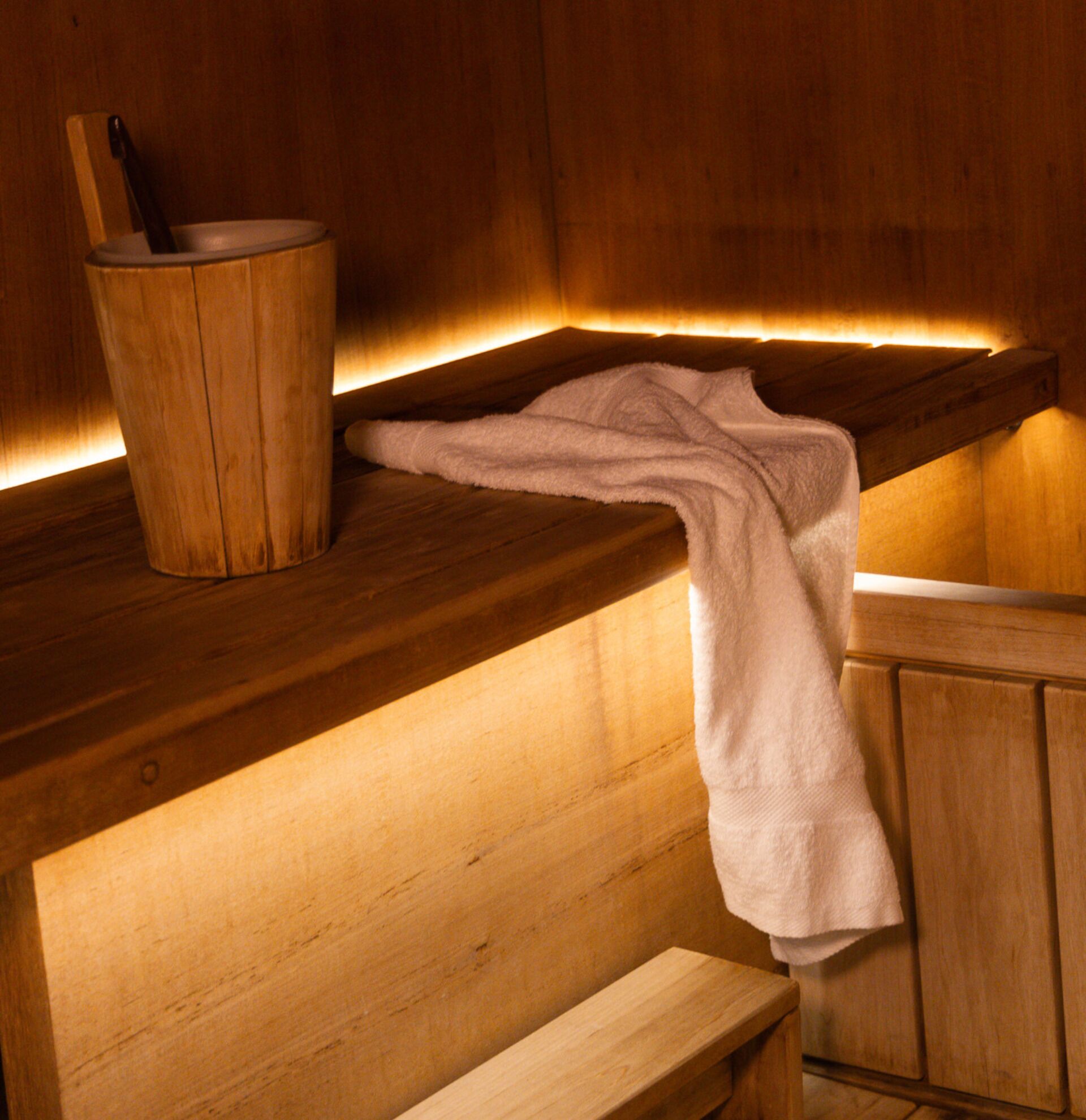 Eine hölzerne Bank mit einem darübergelegten Handtuch, eine Holzschale mit einem Löffel und eine Holzwand mit einer warmen Lichtleiste.
