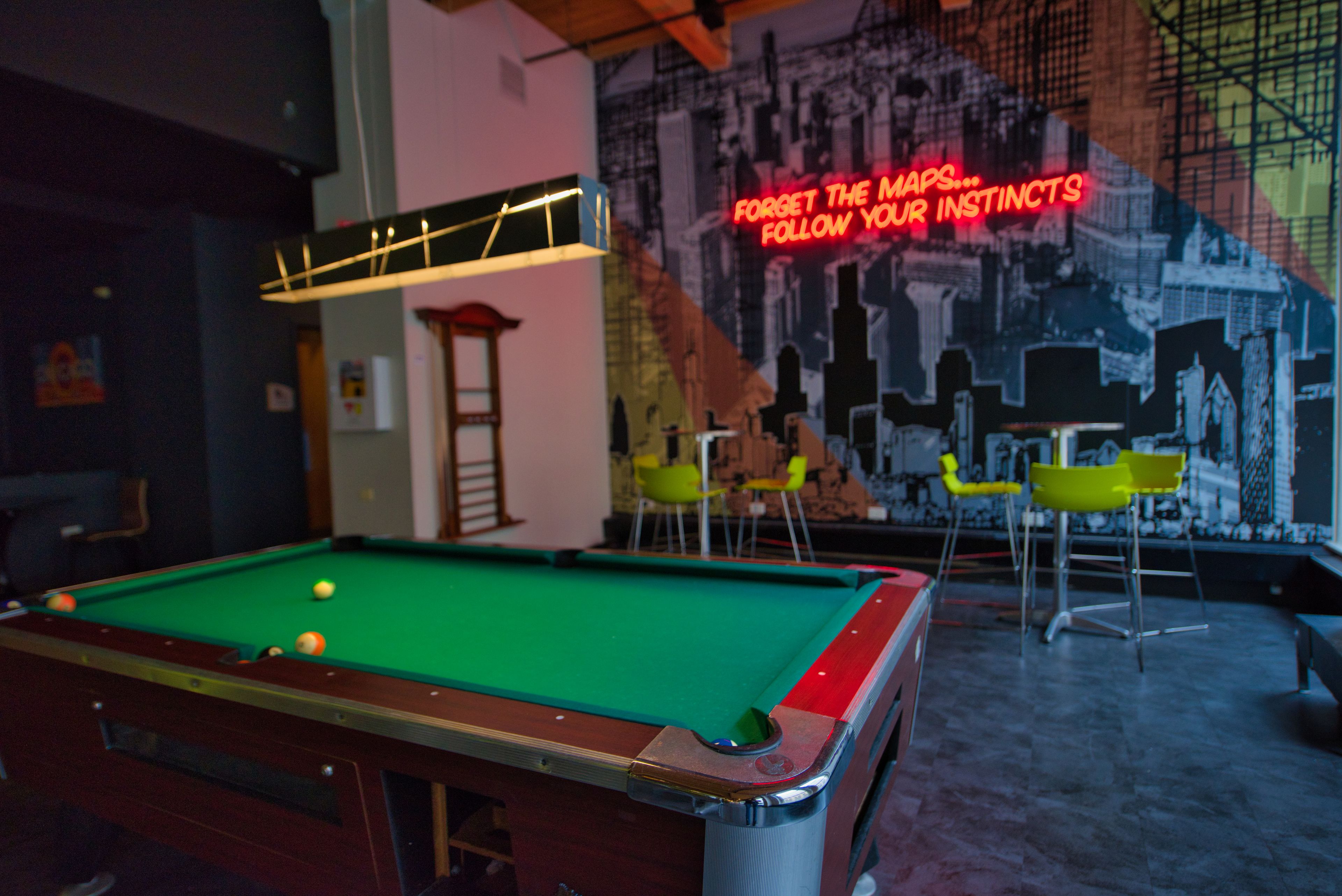 A pool table with balls, a neon sign, and yellow chairs in a room with a cityscape mural.
