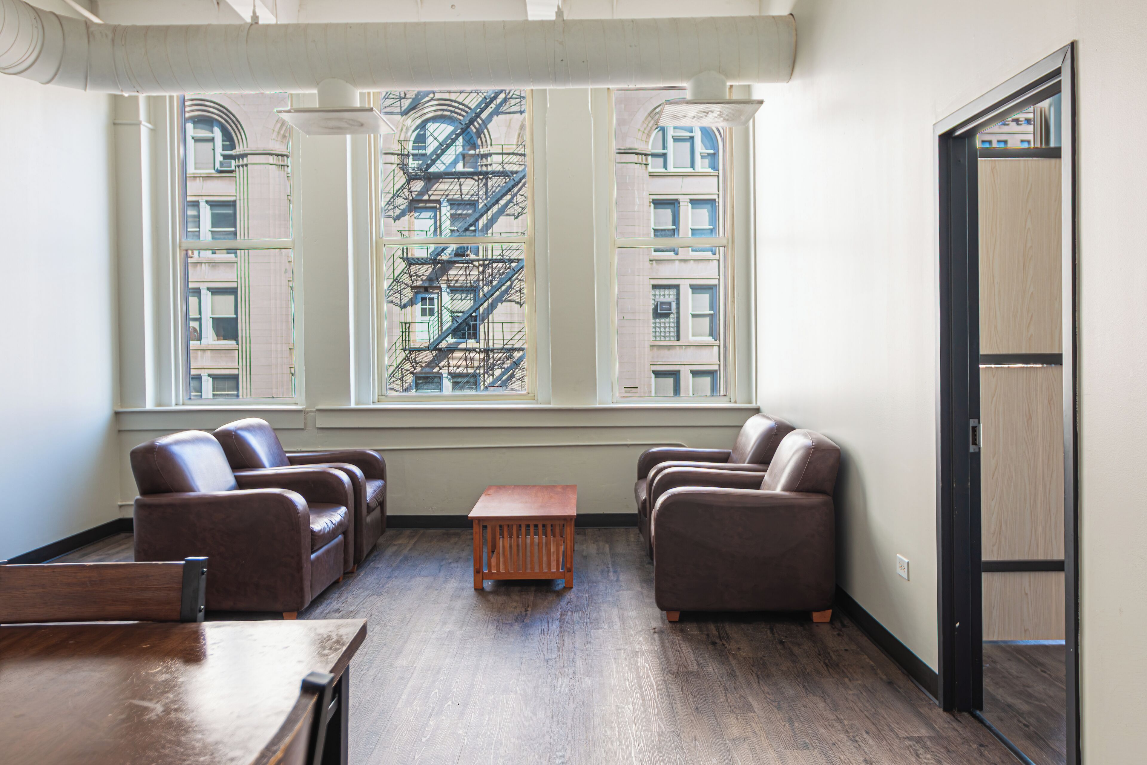 A spacious room with a large window, two leather sofas, a wooden coffee table, and a wooden desk.