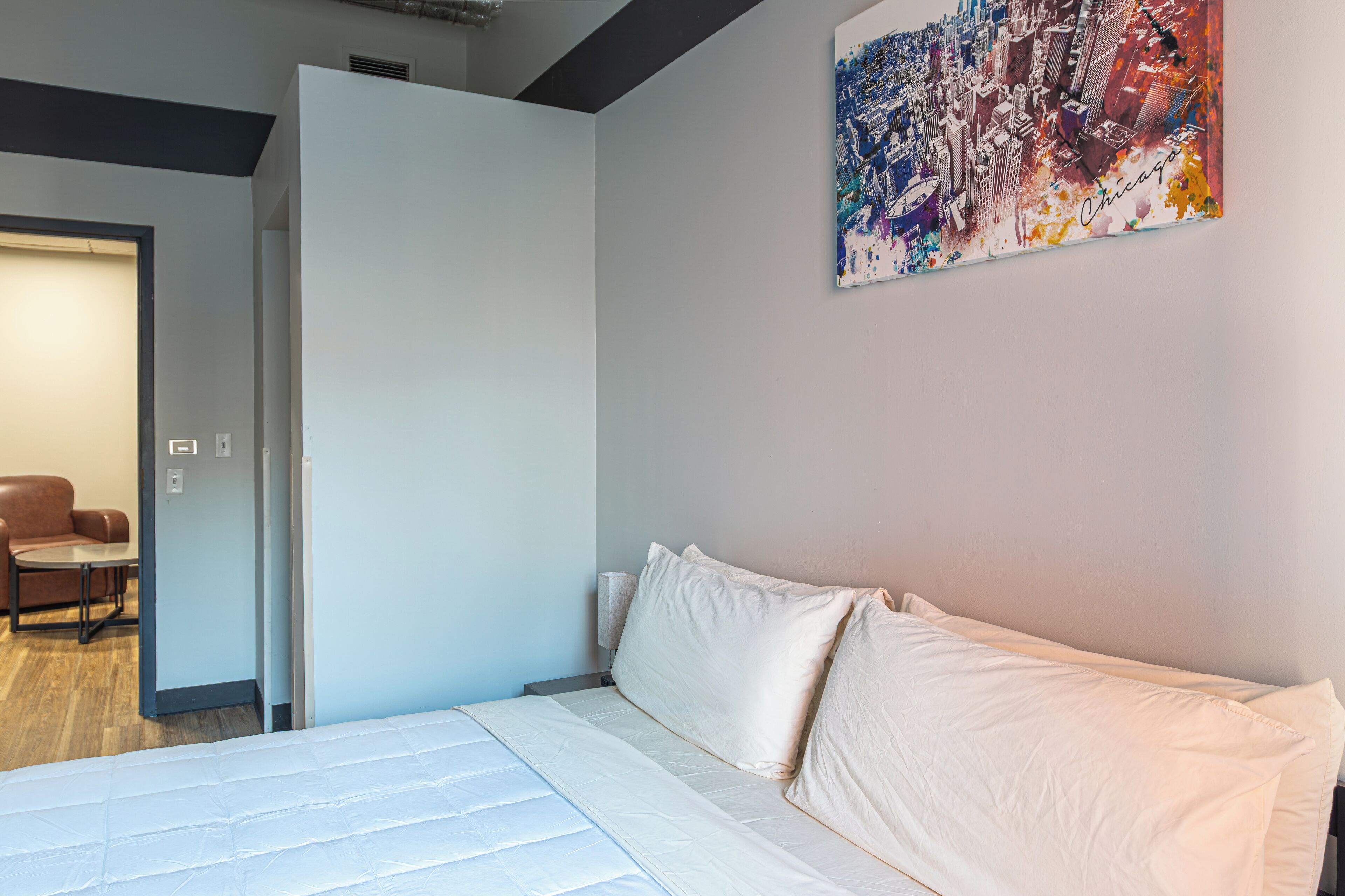 A modern hotel room with a bed, a door leading to a sitting area, and a cityscape painting on the wall.