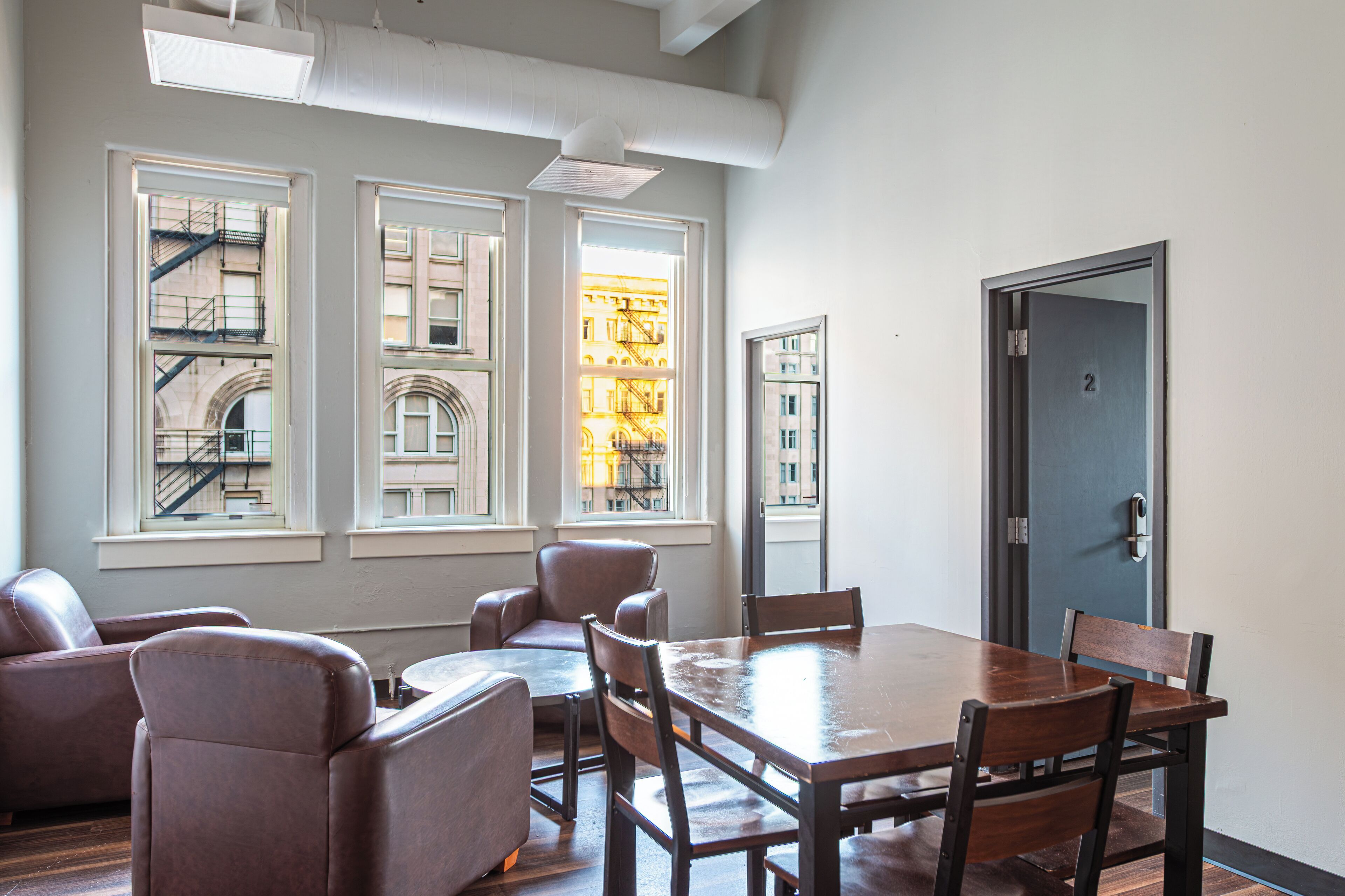 A modern interior with a large wooden table and leather chairs, large windows with views of buildings, and a door with a number.