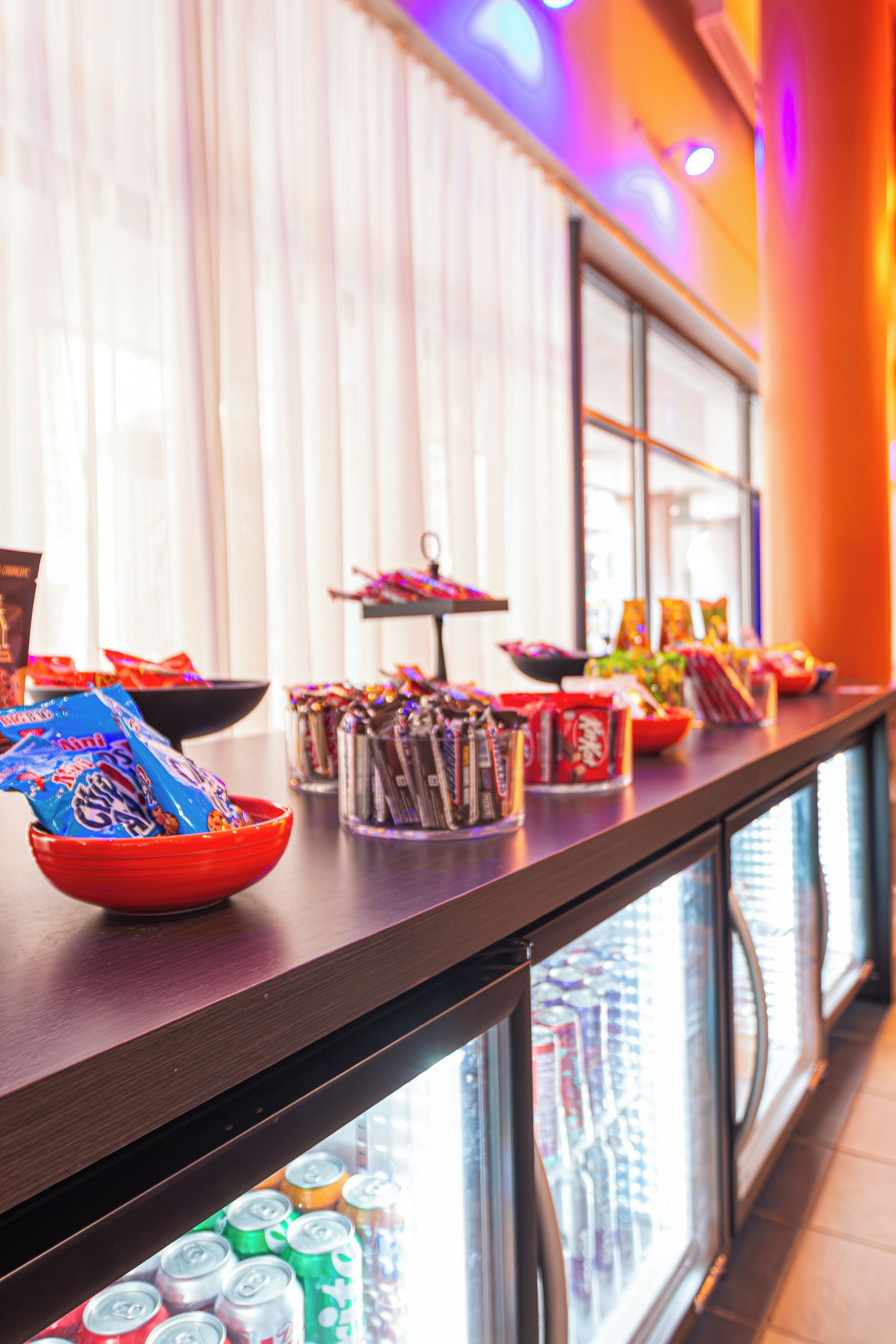 Una mesa de buffet con diversos alimentos y aperitivos, una vitrina con botellas, y un mostrador refrigerado con bebidas.