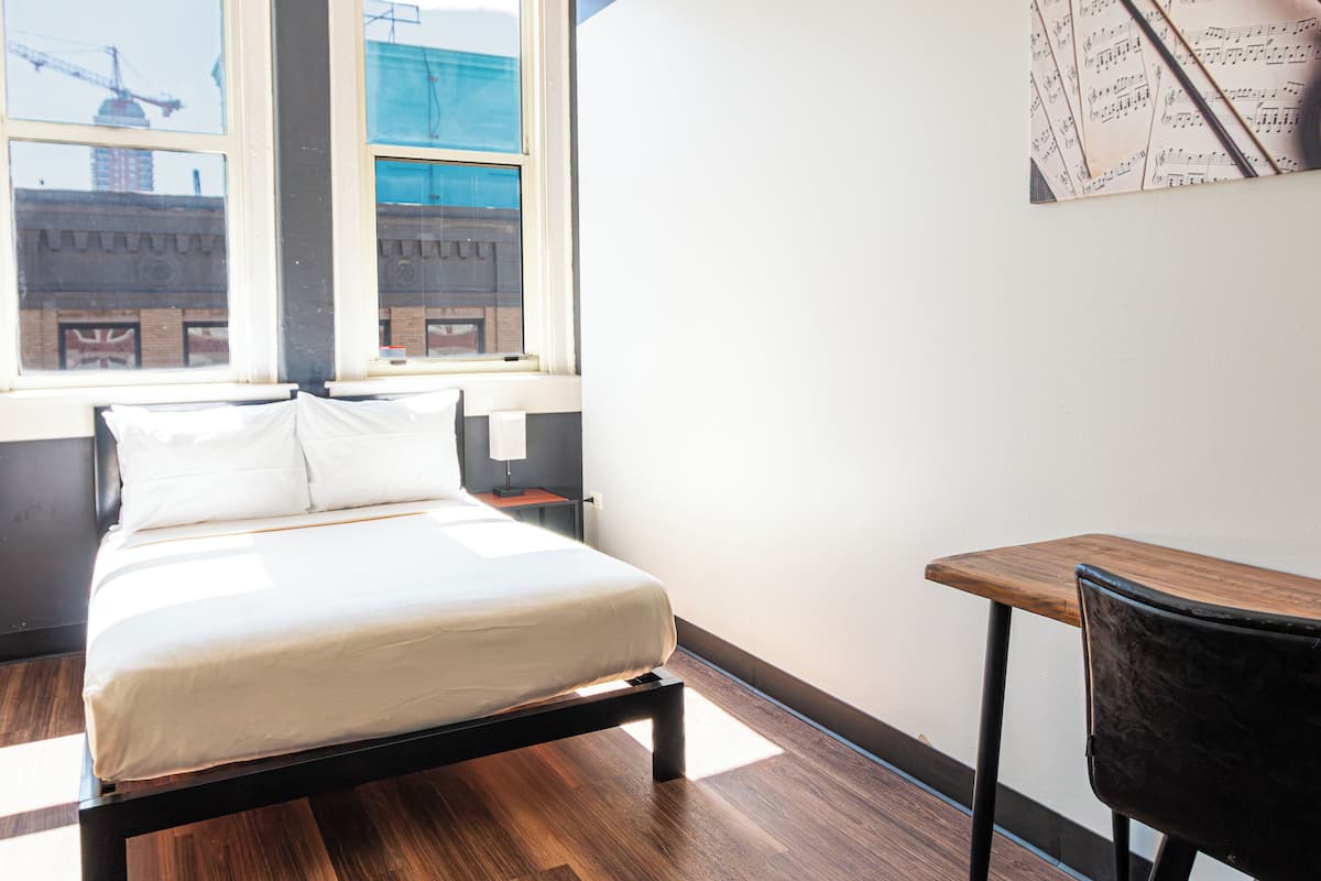 A bedroom with a bed, a desk, and a chair. There is a window with a view of buildings and a crane.