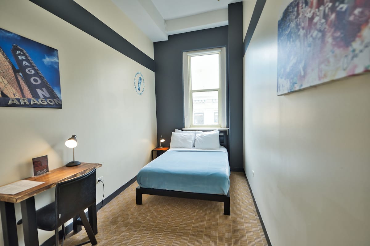 A bedroom with a bed, a desk, a chair, a lamp, and a window.