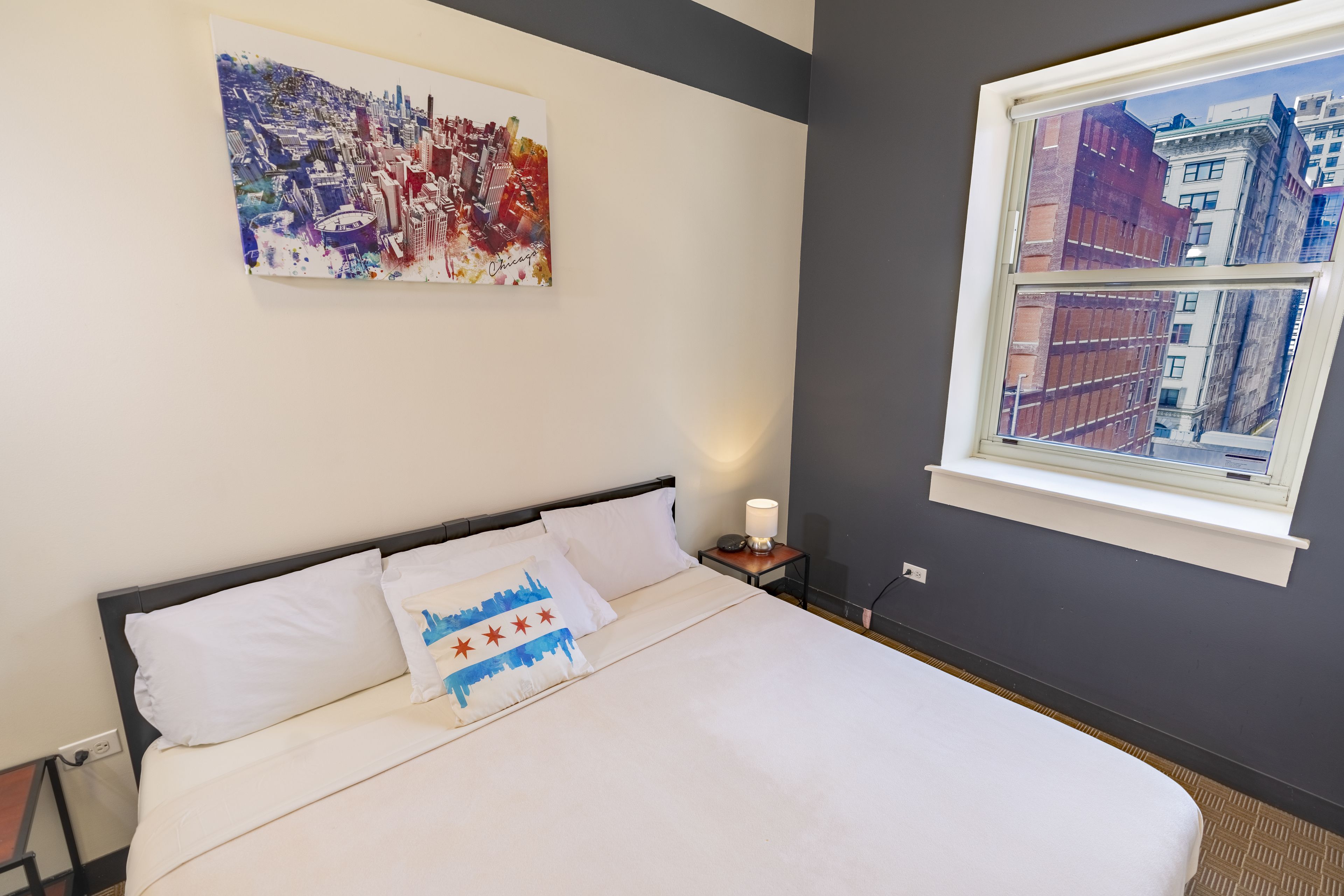 Un lit bien fait, avec du linge de lit blanc et un oreiller orné d’un motif représentant une vue panoramique de la ville. Une peinture abstraite aux couleurs vives est accrochée au-dessus du lit, et une fenêtre donnant sur des immeubles est visible.