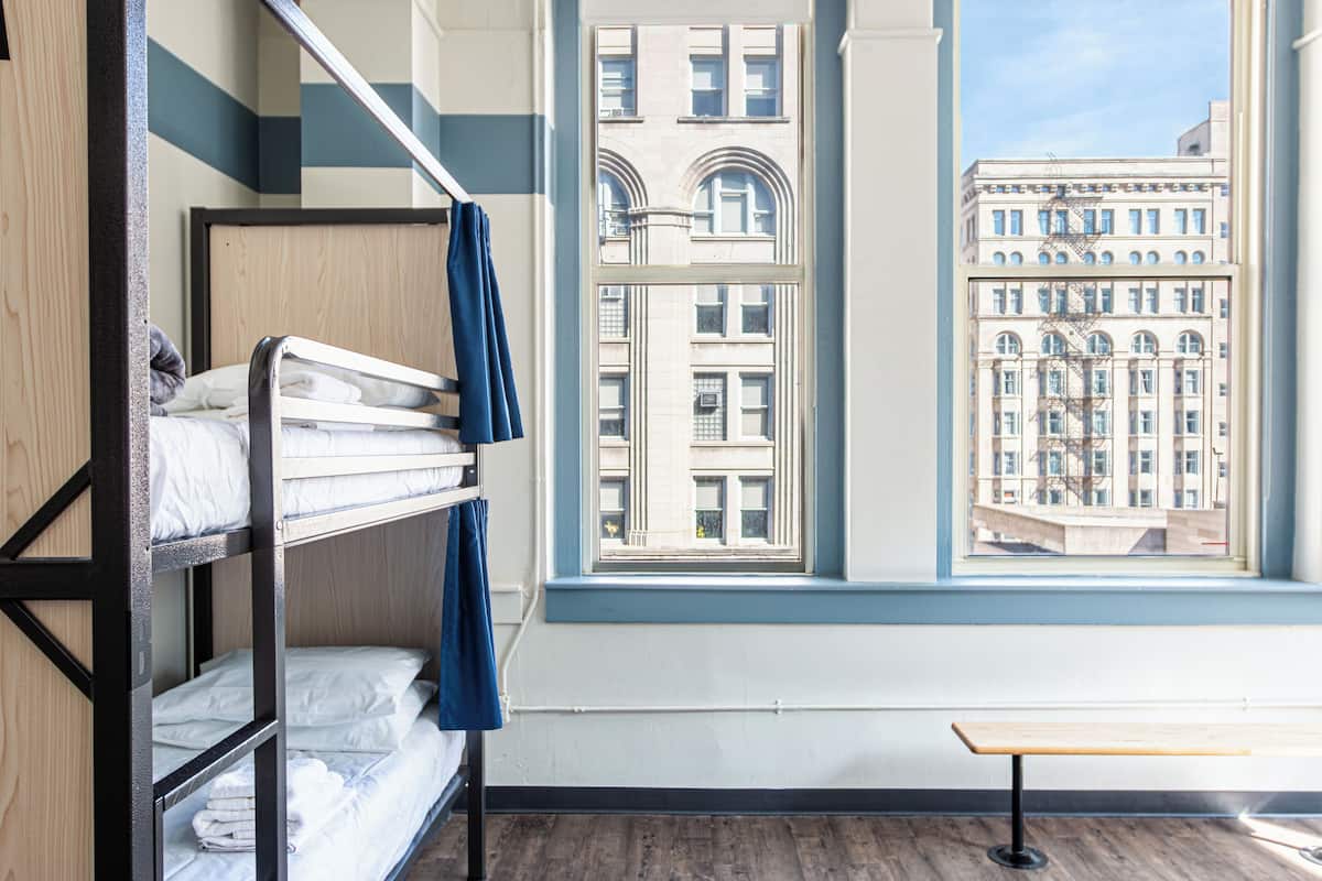 A hotel room with bunk beds, a wooden bench, and a large window offering a view of a historic building.