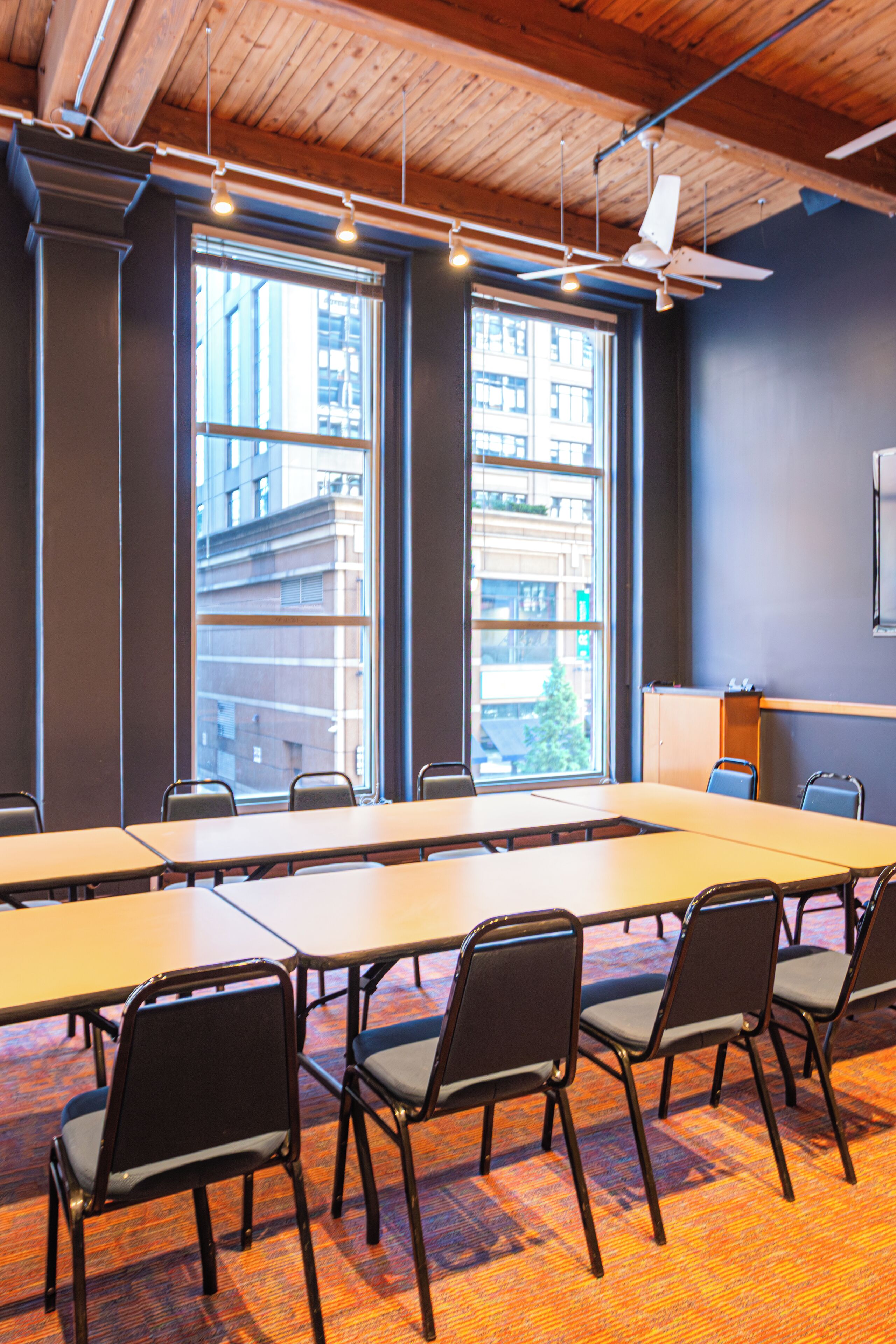 Un salón de conferencias con una larga mesa, varias sillas, ventanas amplias que dan a una vista de la ciudad, un ventilador de techo y una pantalla de proyector.