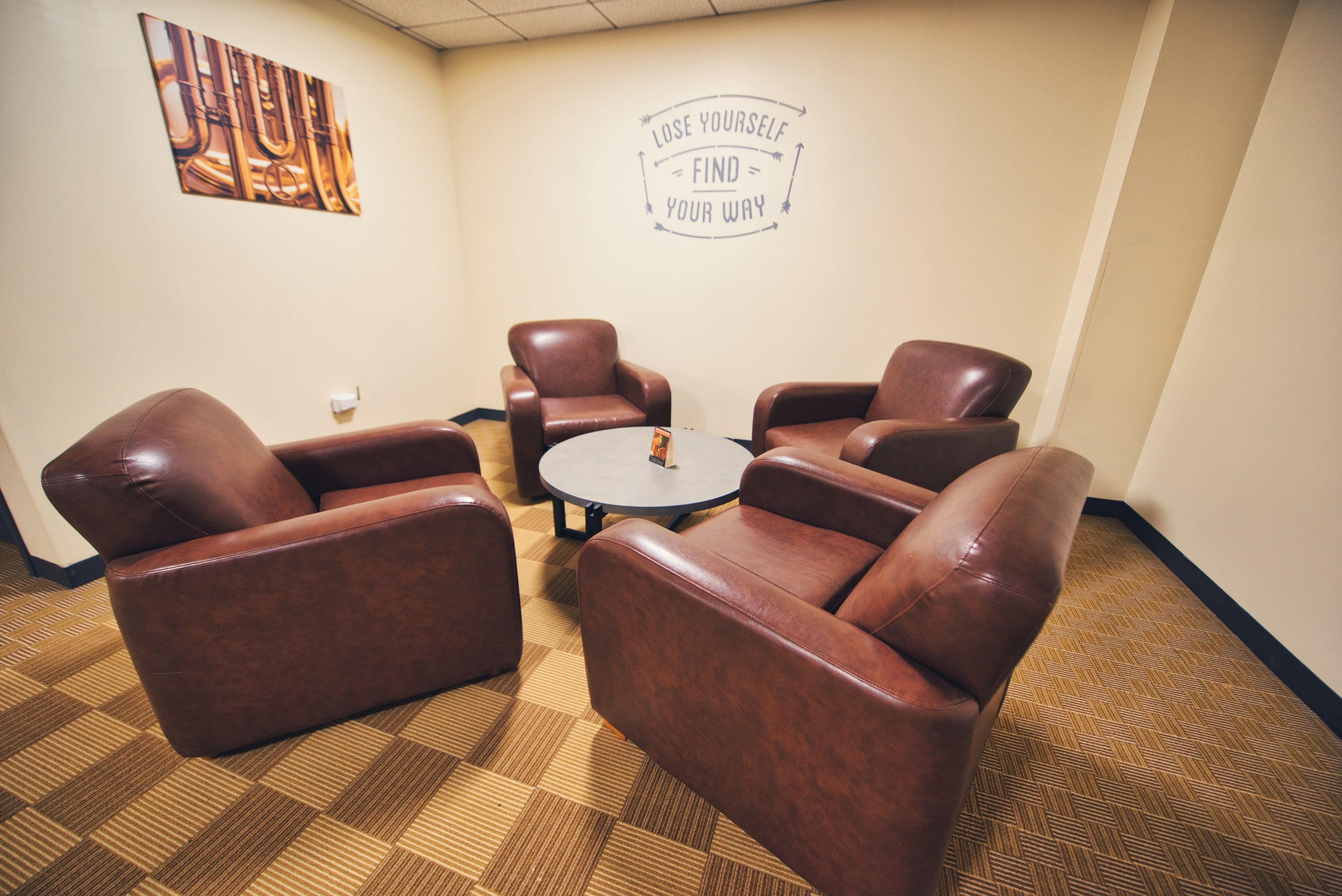 A conference room with four brown leather chairs around a small round table, a motivational quote on the wall, and a patterned carpet.