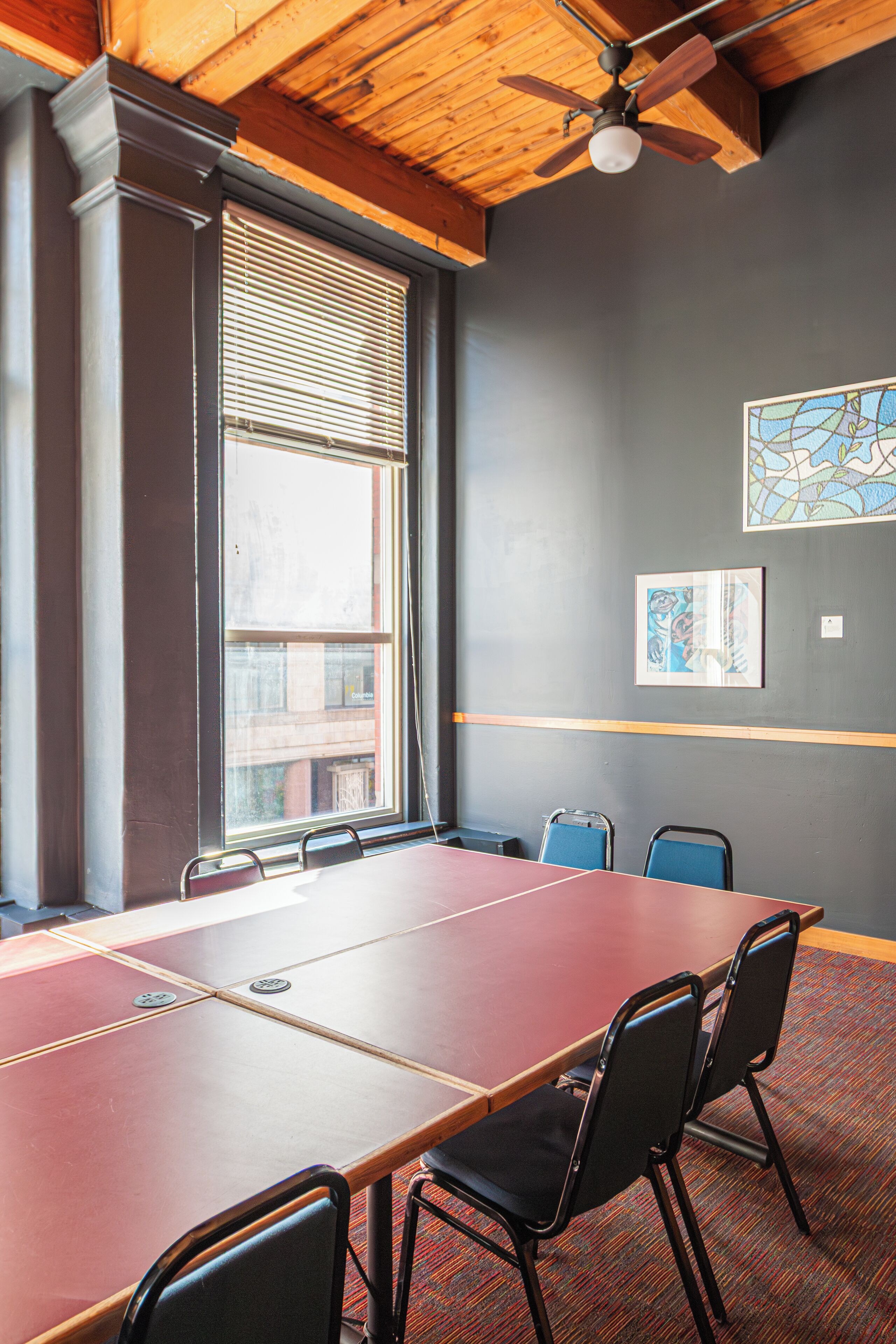 Uma sala de conferências com uma mesa comprida, quatro cadeiras, um ventilador de teto e janelas amplas com persianas.