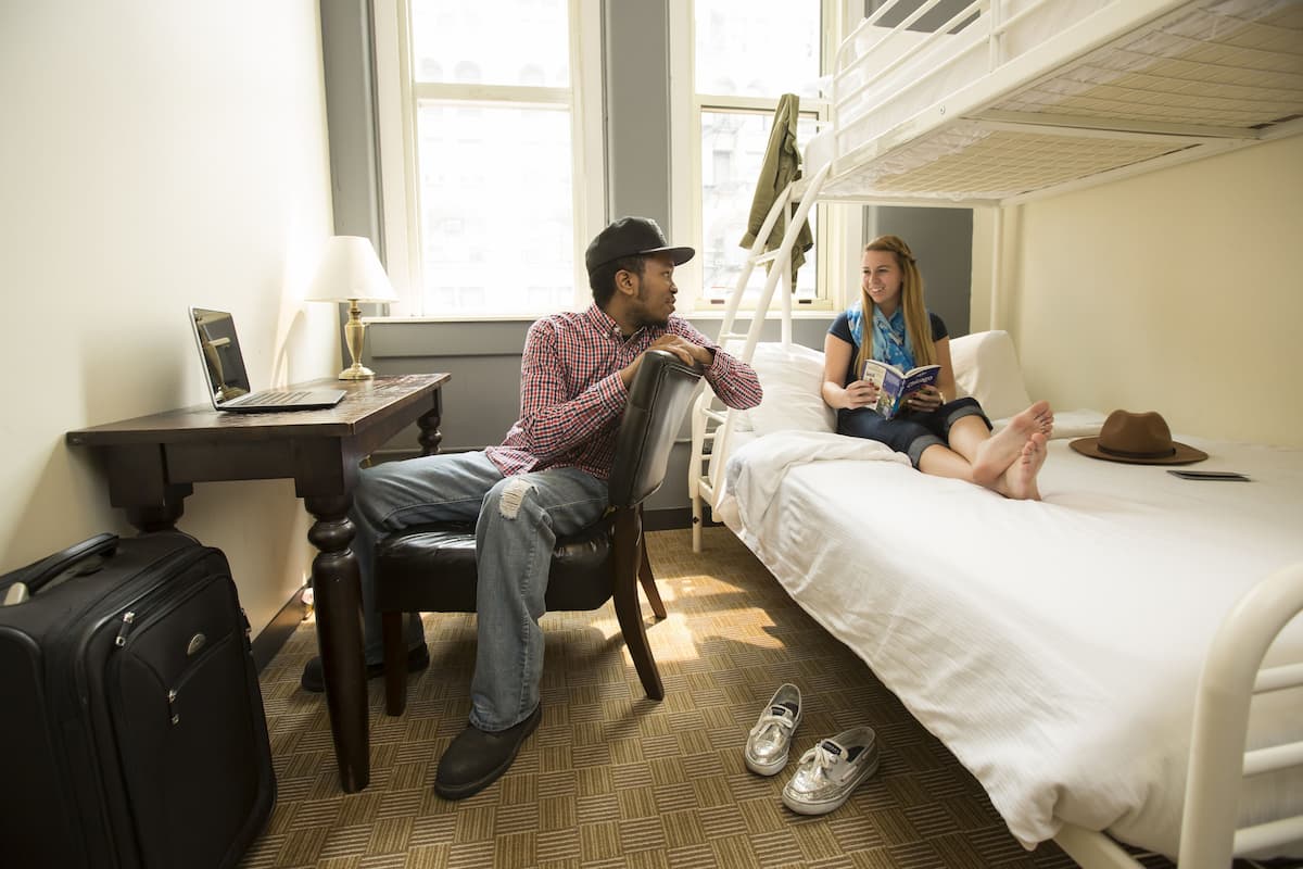 A hotel room with a bed, a desk with a laptop, a suitcase, and a pair of shoes on the floor.