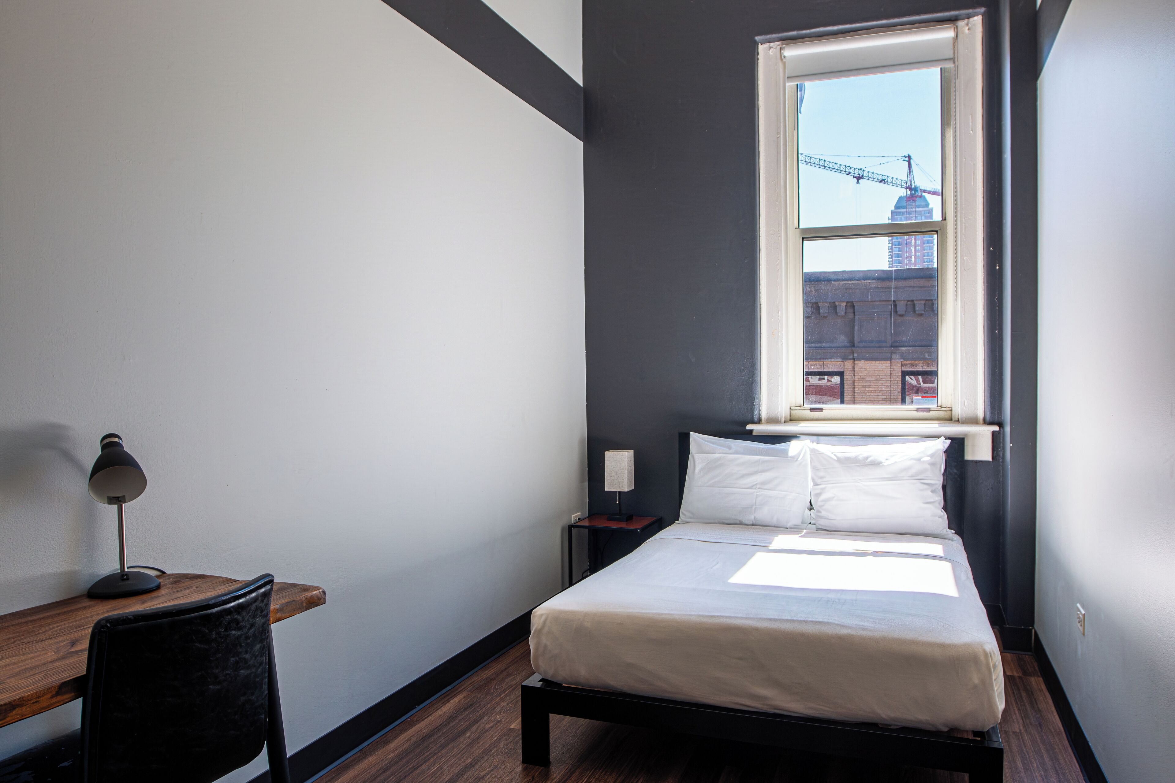 A bedroom with a bed, a desk, a chair, a nightstand, a lamp, and a window with a view of a building and a crane.