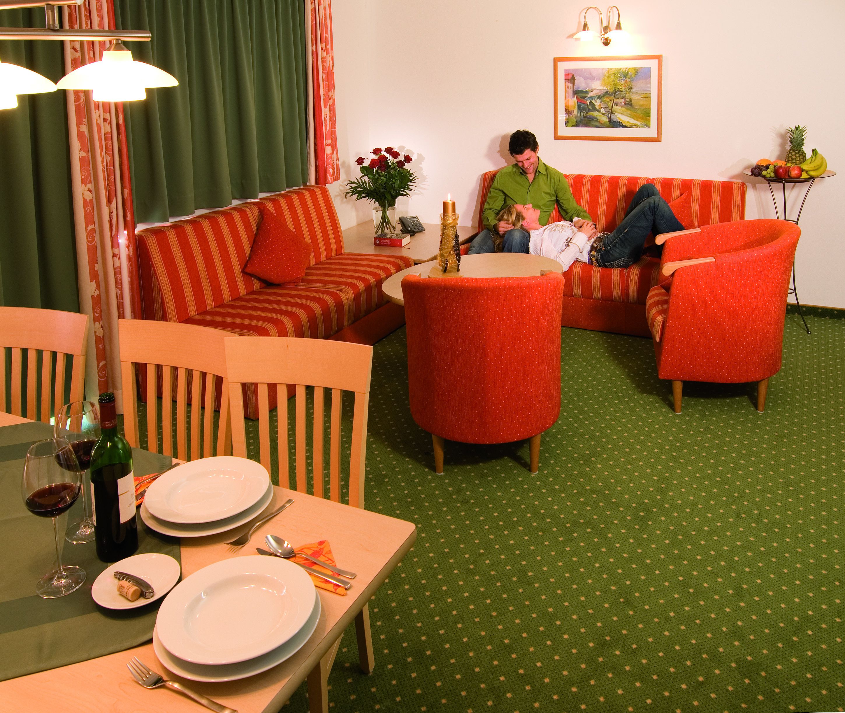 A cozy living room with a sofa, two armchairs, a dining table set with plates and wine glasses, and a small table with a candle and fruit.
