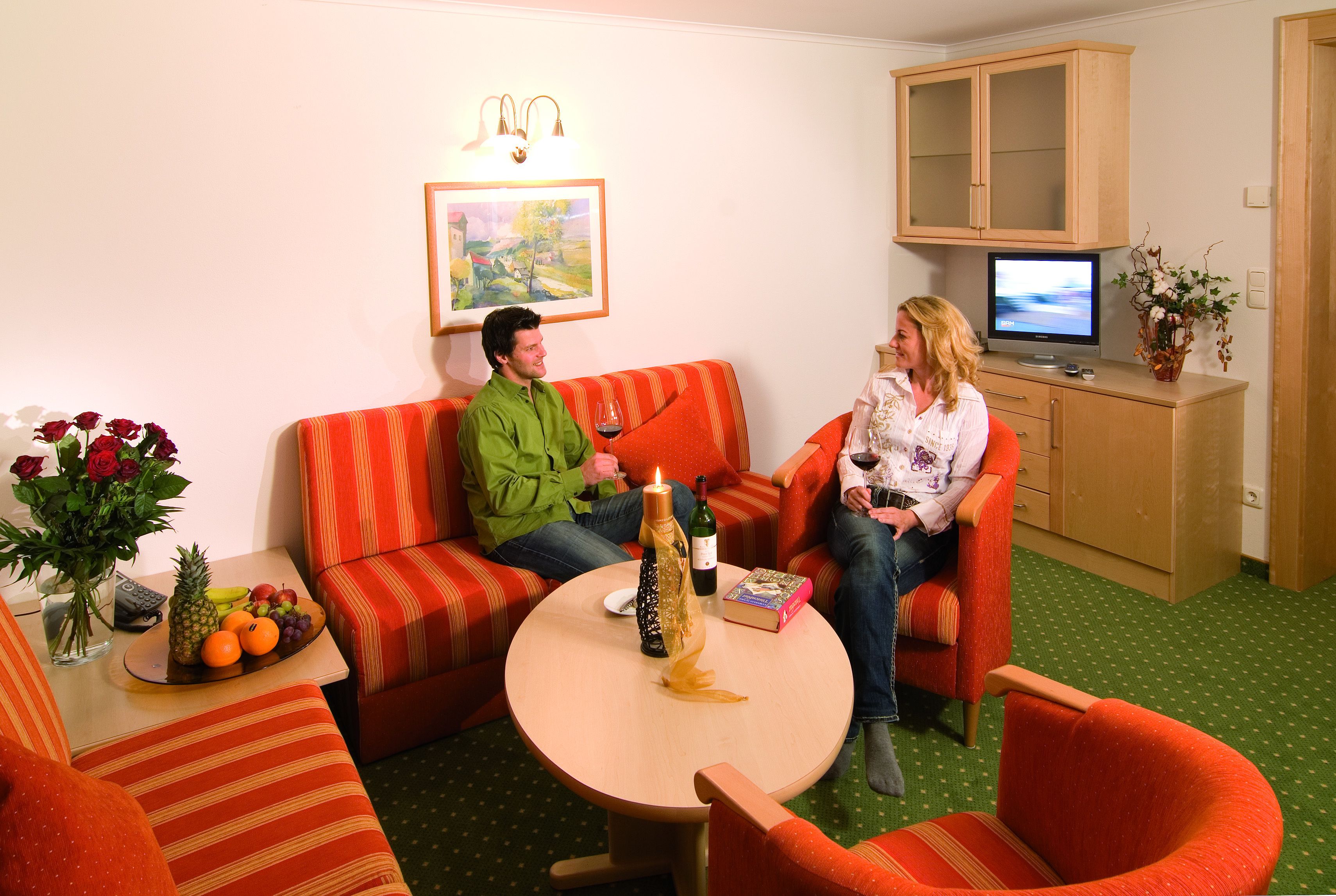 A cozy living room with striped sofas, a small round table with a bottle and a book, and a television on a cabinet.