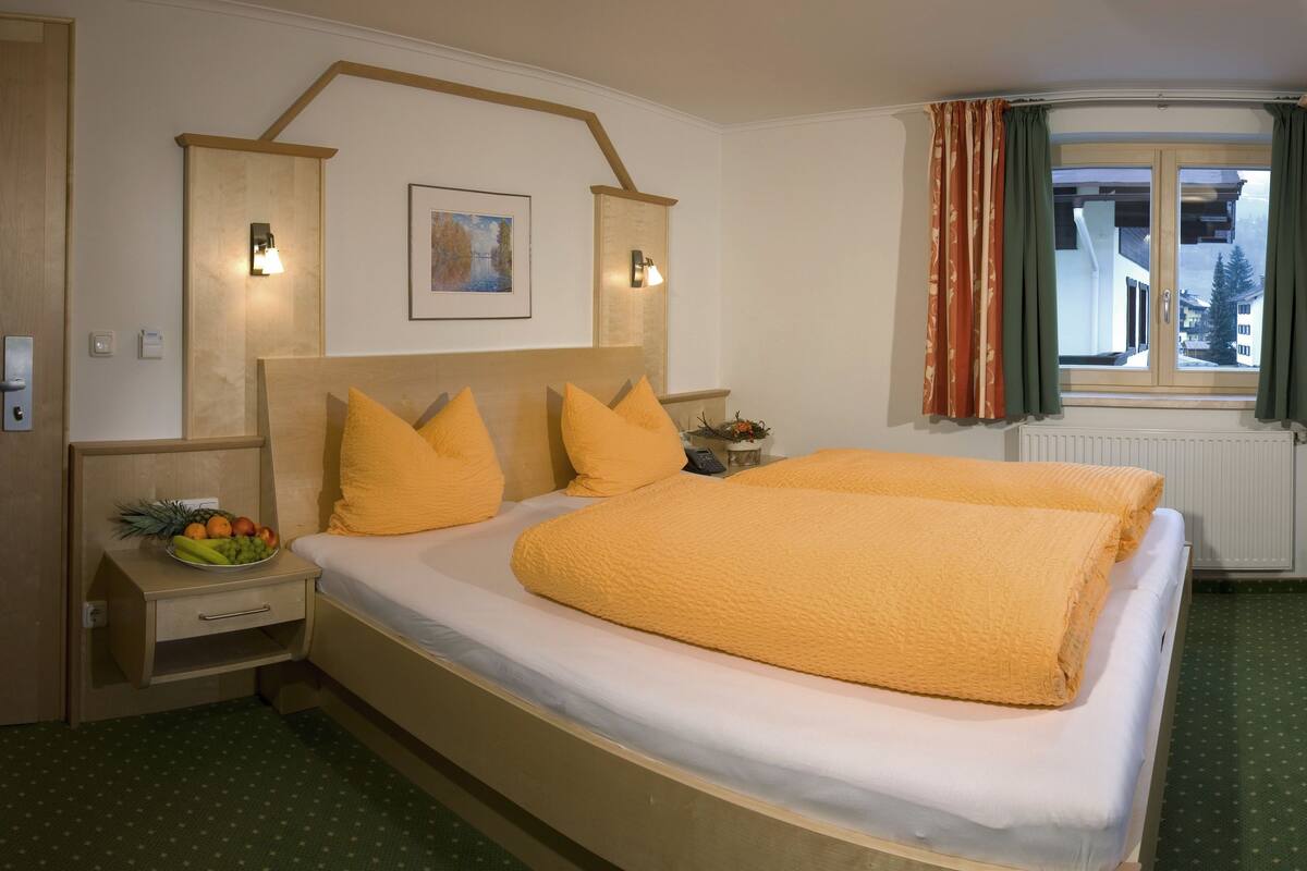 A hotel room with a large bed, a desk, a chair, a window with curtains, and a wall-mounted light.