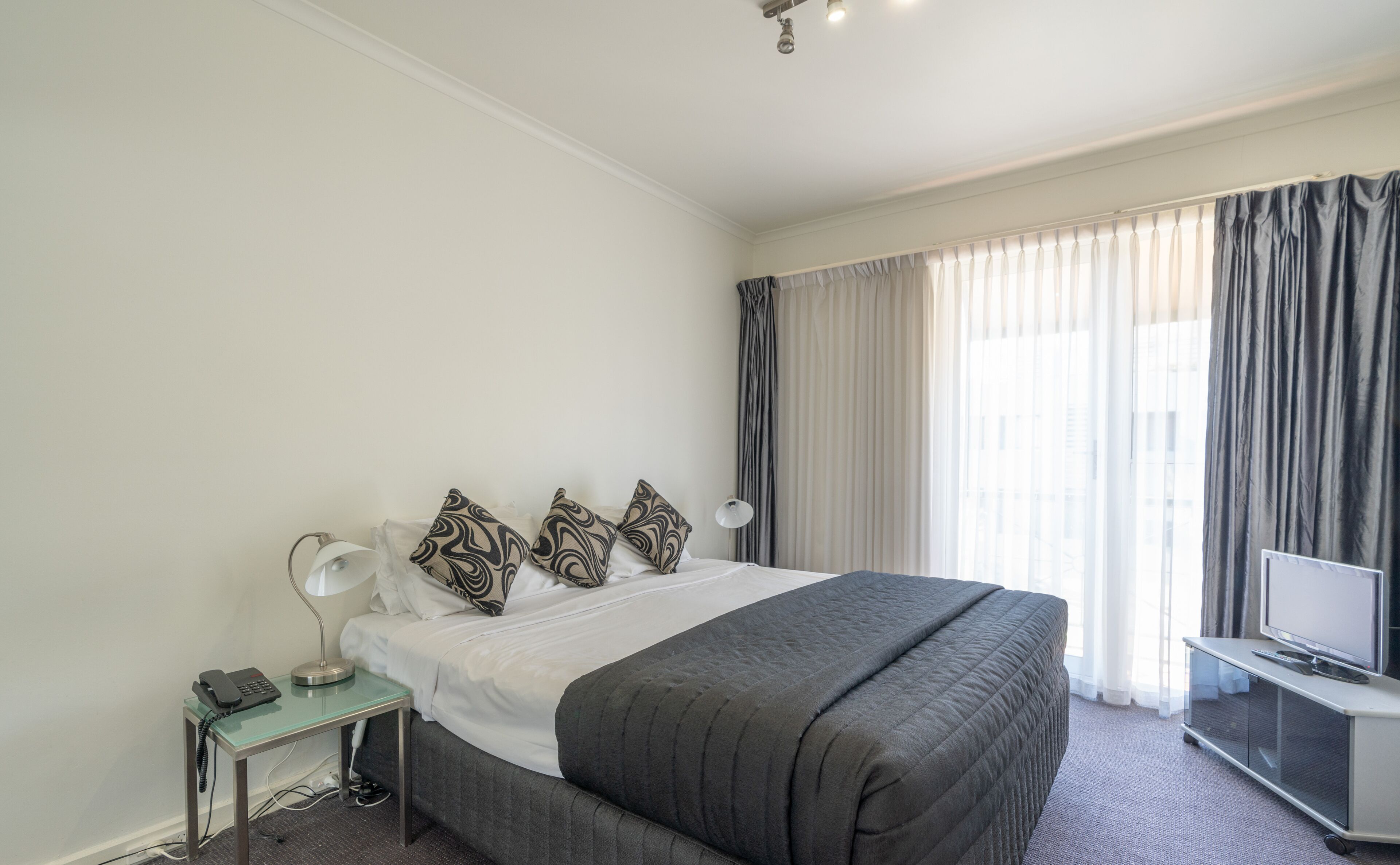 A hotel room with a large bed, a bedside table with a lamp, a TV, and a telephone.