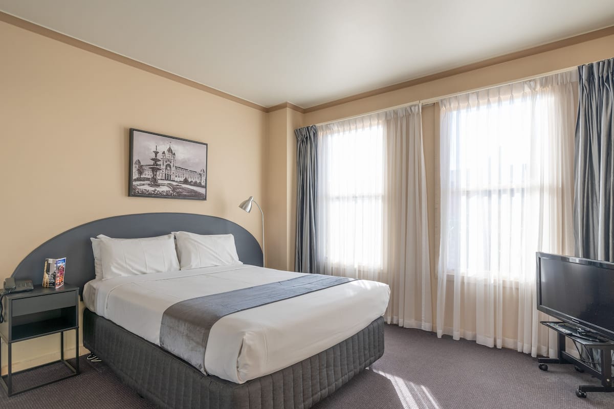 Habitación de hotel con una cama grande, una mesita de noche con teléfono, un televisor sobre un soporte y una ventana con cortinas.
