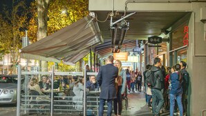 A bustling outdoor café with tables and chairs under a covered area, surrounded by trees and street signs.