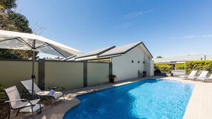 Una piscina con sillones de descanso y sombrillas, rodeada por un muro bajo y un edificio con techo plano.