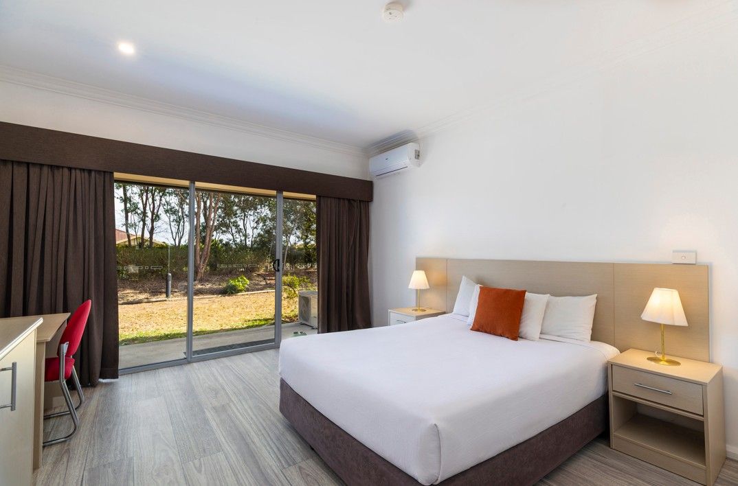 Una habitación de hotel con una cama grande, dos mesitas de noche con lámparas, un escritorio con una silla y un ventanal corredero que da a un espacio exterior.
