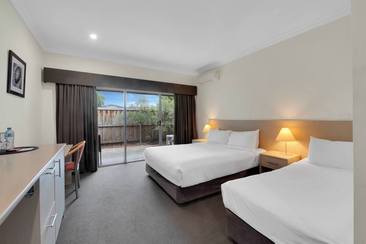 A hotel room with two beds, a desk, a chair, and a view of a wooden fence and trees.