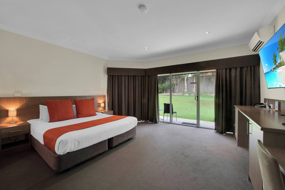 A hotel room with a large bed, a desk, a chair, a TV, and a sliding glass door to a garden.
