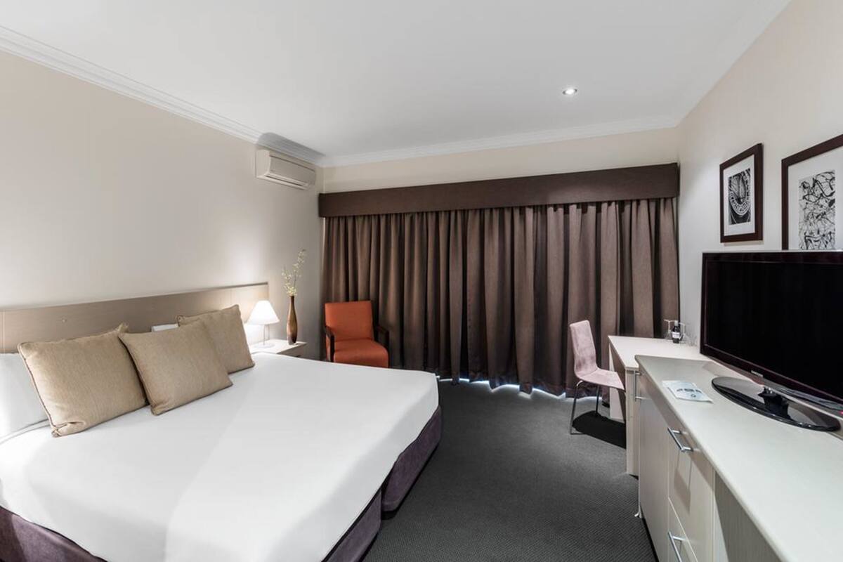 A hotel room with a large bed, a desk with a flat-screen TV, a chair, a desk lamp, and a window with curtains.