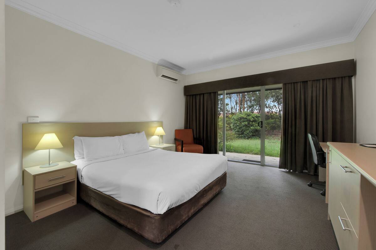 A hotel room with a large bed, bedside lamps, a desk, and a view of the outdoors through a sliding glass door.