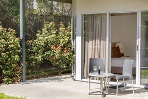 Habitación de hotel moderna con una puerta de vidrio que da a un balcón, una pequeña mesa con una silla y una cama con un almohadón.