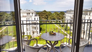 Un balcón con sillas de mimbre y una mesa, con vistas a un jardín cuidado y edificios.