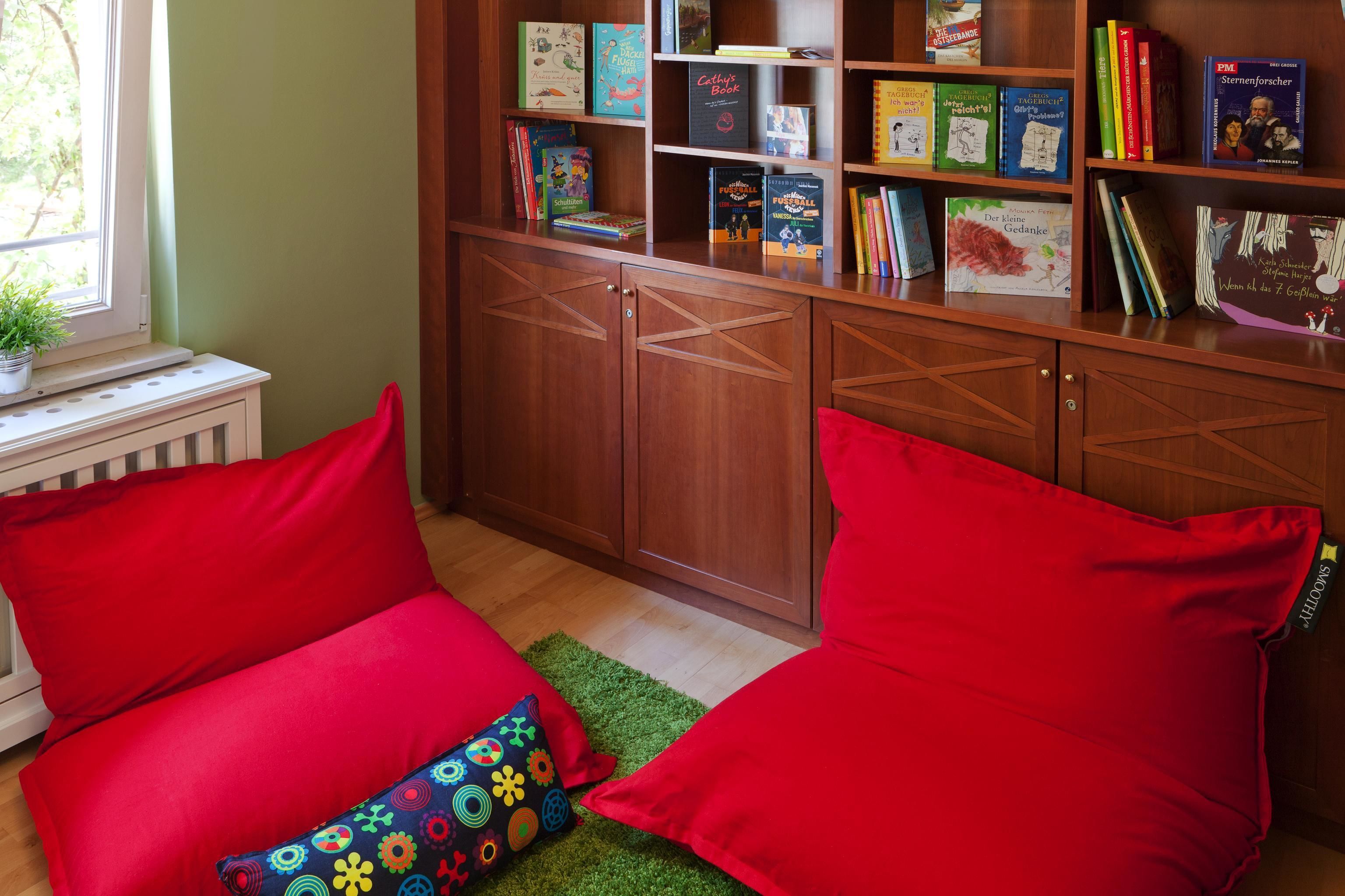 A cozy reading nook with red cushions, a bookshelf filled with books, and a window with a plant.