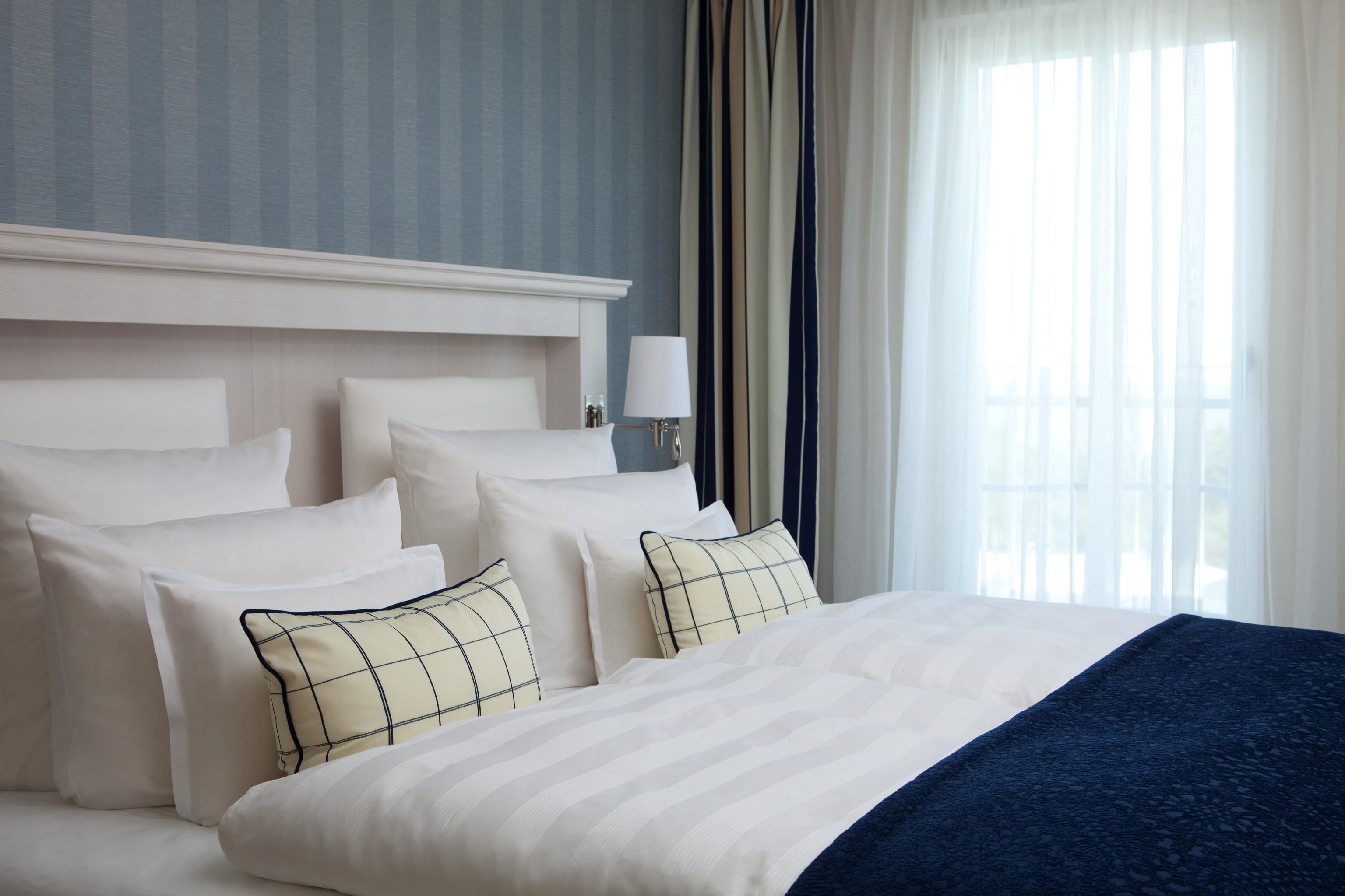Uma cama bem arrumada com lençóis brancos e um cobertor azul, um abajur e vista para uma janela com cortinas leves.