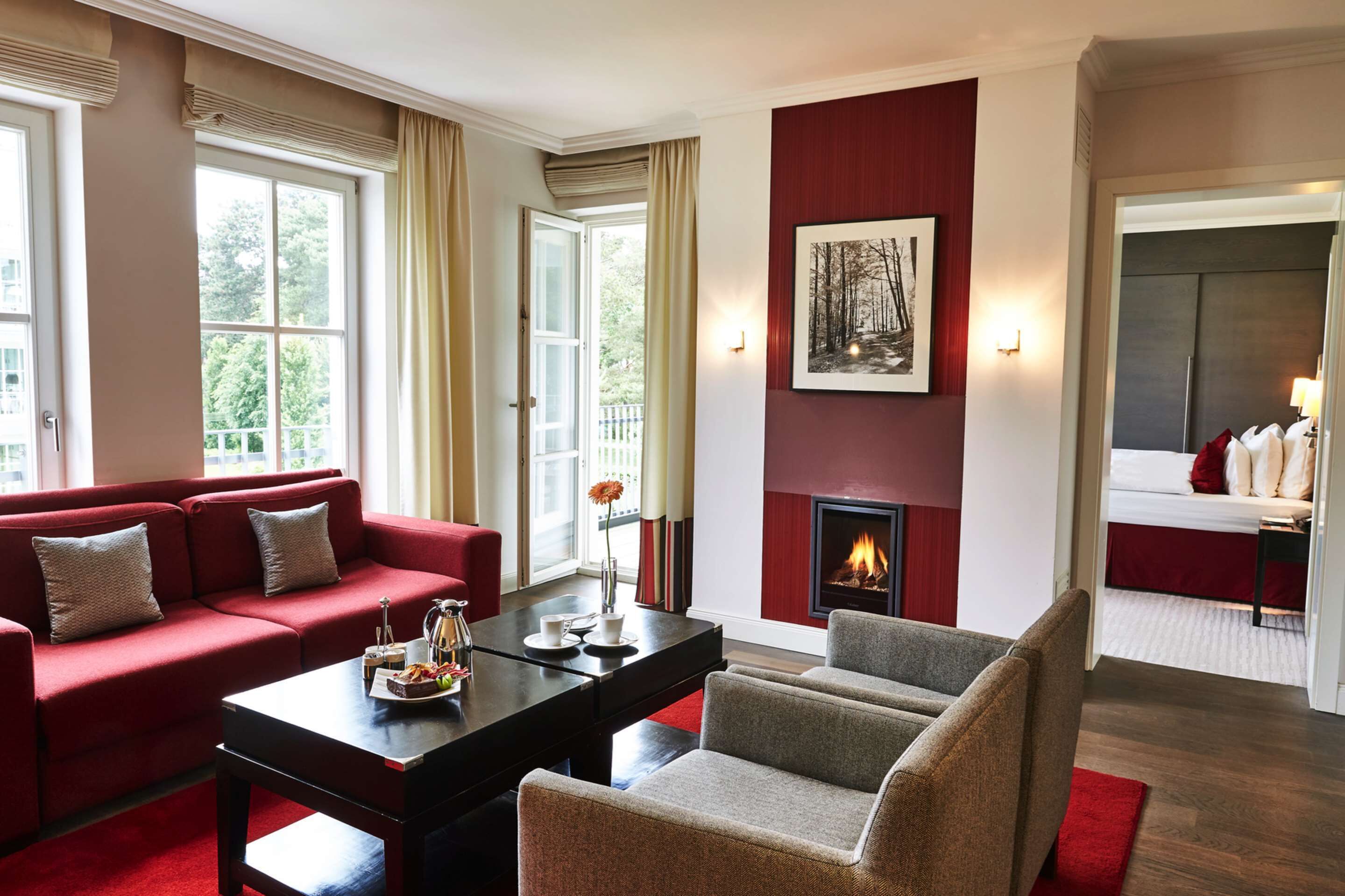 Un salon moderne avec un canapé rouge, un fauteuil gris et une table basse noire. On y trouve une cheminée surmontée d’un tableau encadré, et un balcon visible à travers les fenêtres.