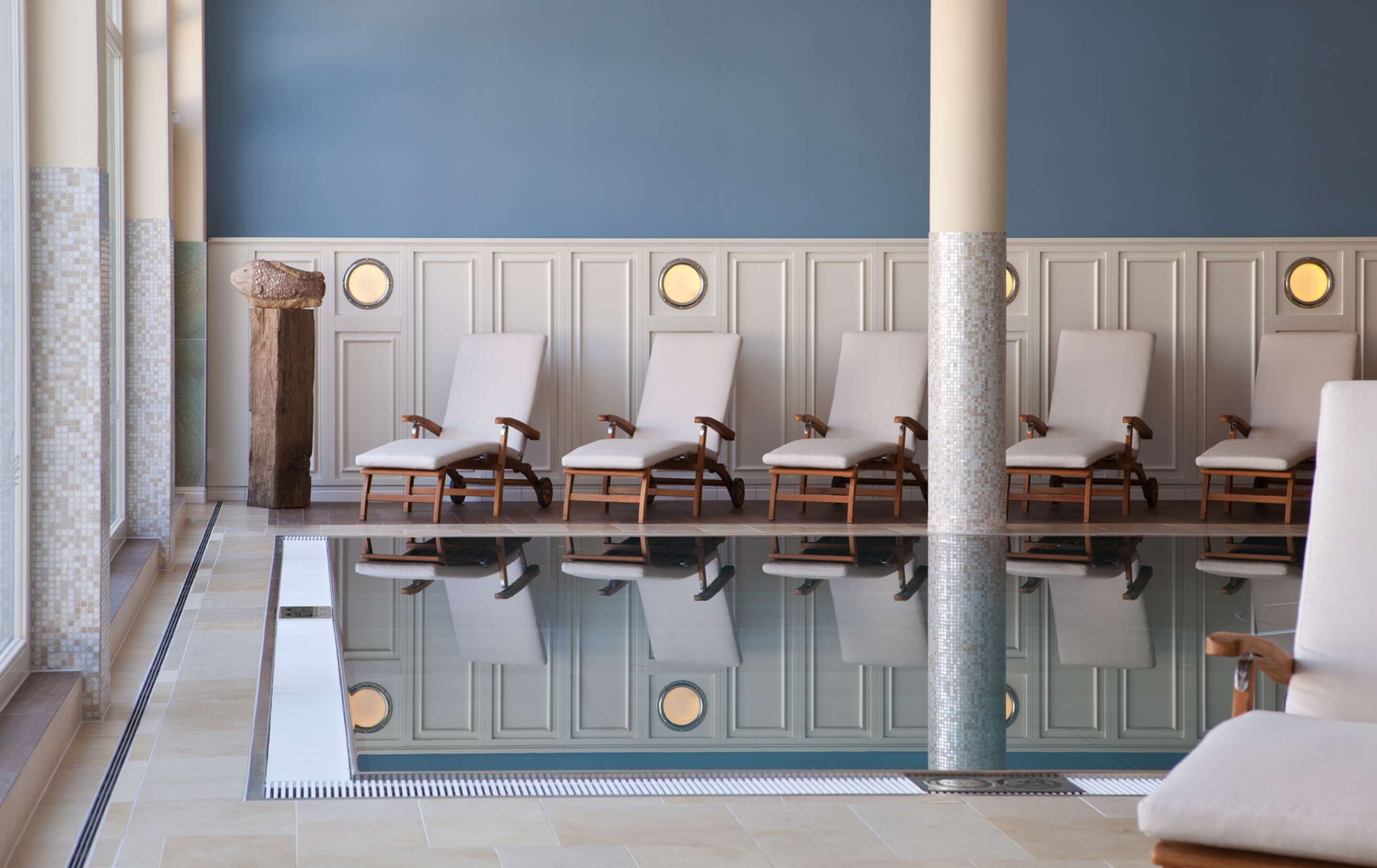 A pool area with six lounge chairs aligned along the poolside, a decorative column, and a tiled floor.