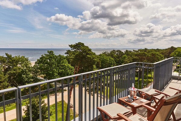A balcony with a view of the sea and trees, wooden furniture, and a table with a bottle and glasses.