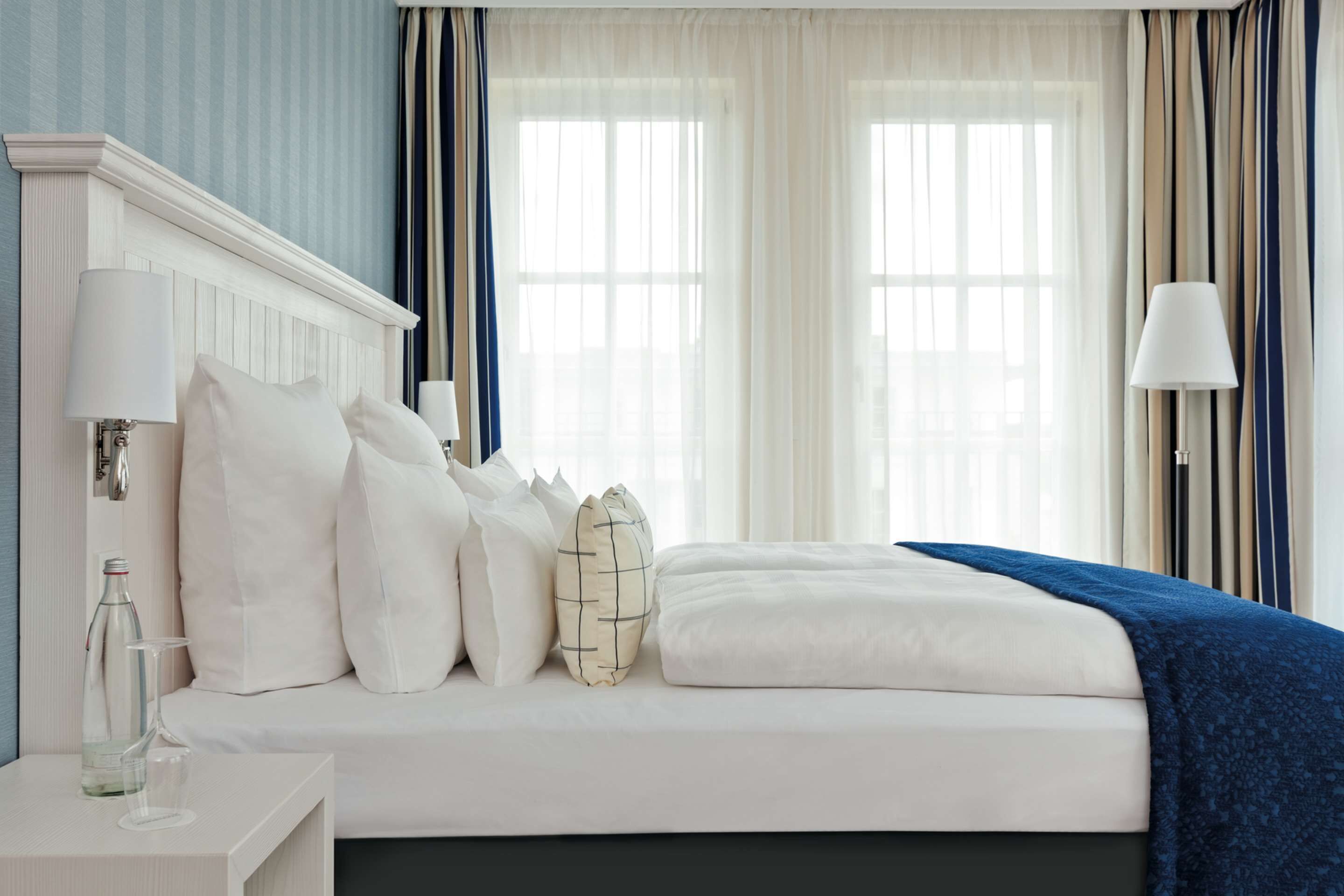 Un lit bien fait, avec du linge de lit blanc et un plaid bleu, une table de chevet avec une lampe, et une fenêtre ornée de rideaux légers.