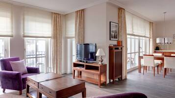 A spacious living room with a flat-screen TV, a wooden coffee table, purple armchairs, and a dining area with a white table and chairs.